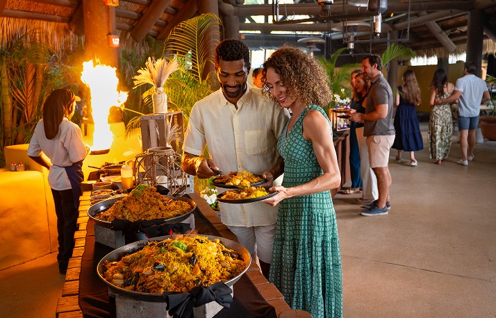 A buffet line under an open sided thatched roof, with a man in a cream linen shirt holding serving tongs and a plate of yellow rice beside a woman in a green patterned dress holding a matching plate, three wide shallow pans of rice dishes along the counter and a tall flame burning behind them