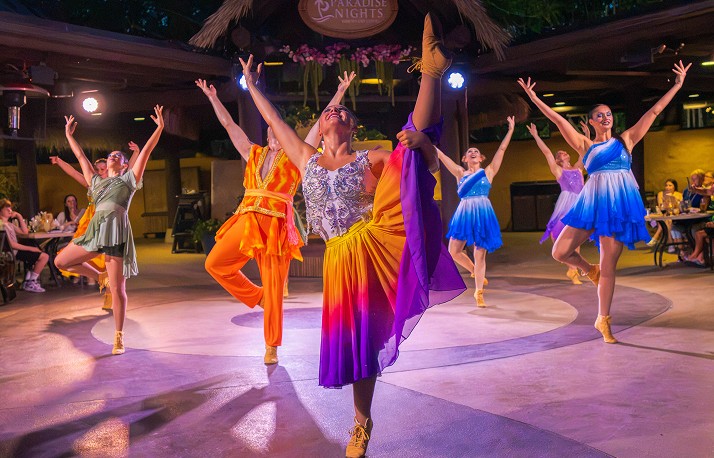A troupe of dancers frozen mid pose with arms raised on a circular floor under a thatched open air pavilion, the lead dancer in an orange to purple ombre skirt, the others in blue and lilac tutus, a sage green dress and an orange jumpsuit, one leg extended high above the group, a round Paradise Nights sign overhead and seated diners at tables on both sides
