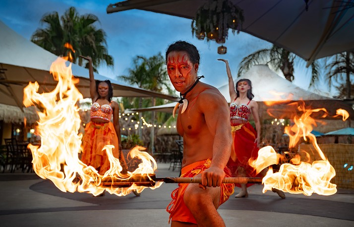 Three fire performers outdoors at dusk, a shirtless male dancer in an orange sarong with red face paint spinning a double ended flaming staff toward the camera and two female dancers in orange and red costumes posing behind him under white shade sails and string lights