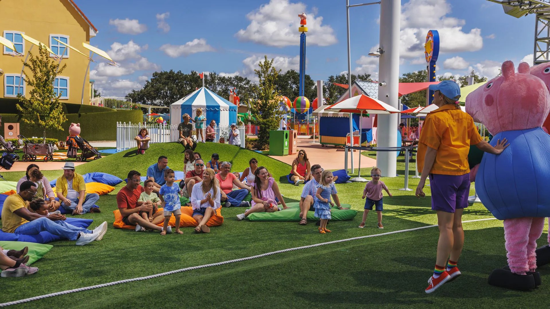 Parents and small children sitting on orange, blue and green floor cushions on artificial turf watching two costumed Peppa Pig characters and a park host, with a striped tent and the yellow Peppa house behind them under a partly cloudy sky