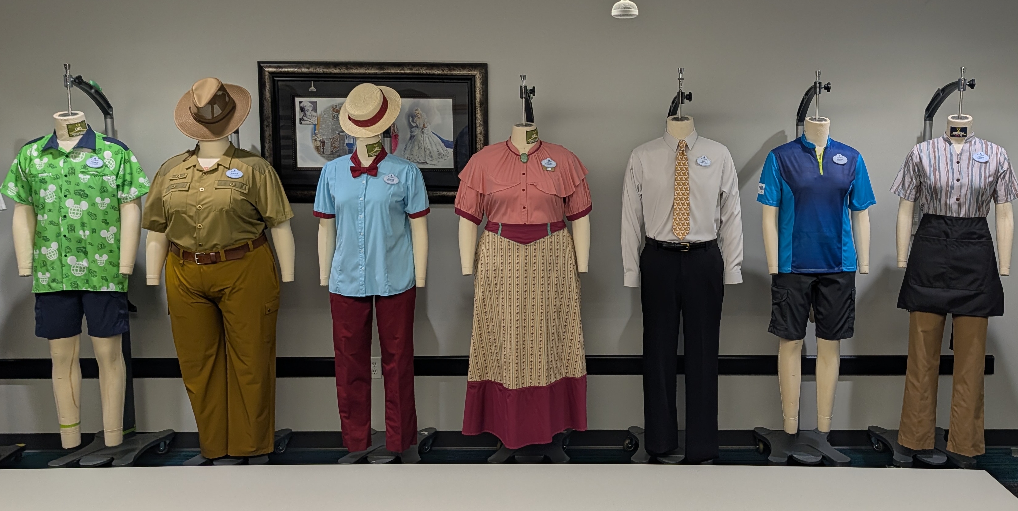 Seven costumes on headless mannequins lined up against a grey wall, including the green parking shirt with navy shorts, an olive Jungle Cruise skipper outfit with a wide brimmed hat, a light blue blouse with a straw boater, a long patterned skirt with a pink ruffled top, a grey dress shirt with a gold necktie, a blue polo with black cargo shorts, and a striped blouse with a black apron