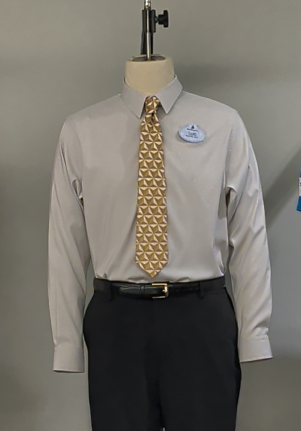 A grey long sleeve dress shirt on a mannequin with black trousers and a black belt, wearing a necktie printed edge to edge with gold triangular Spaceship Earth facets, with an oval Disney name tag on the chest