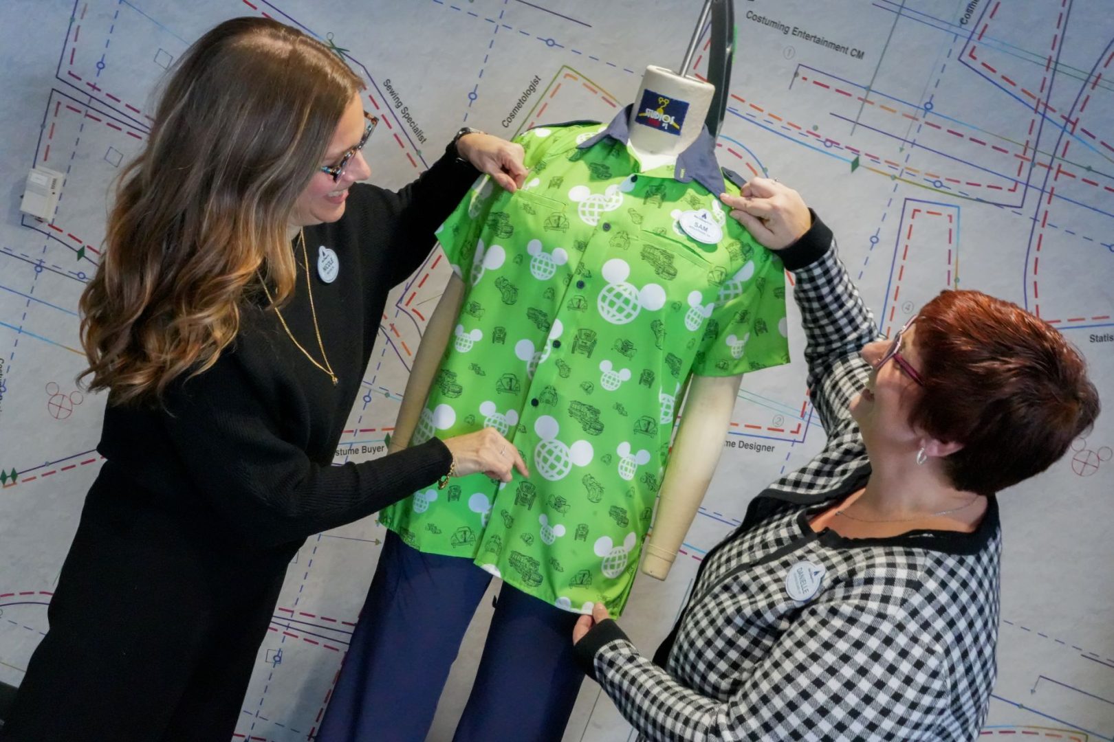 Two Disney Costuming cast members holding up a bright lime green short sleeve button down on a headless dress form; the shirt is printed with white Mickey shaped globes and small dark outline drawings of vehicles, in front of a wall covered in a blueprint style graphic labeled with roles like Costume Buyer and Sewing Specialist