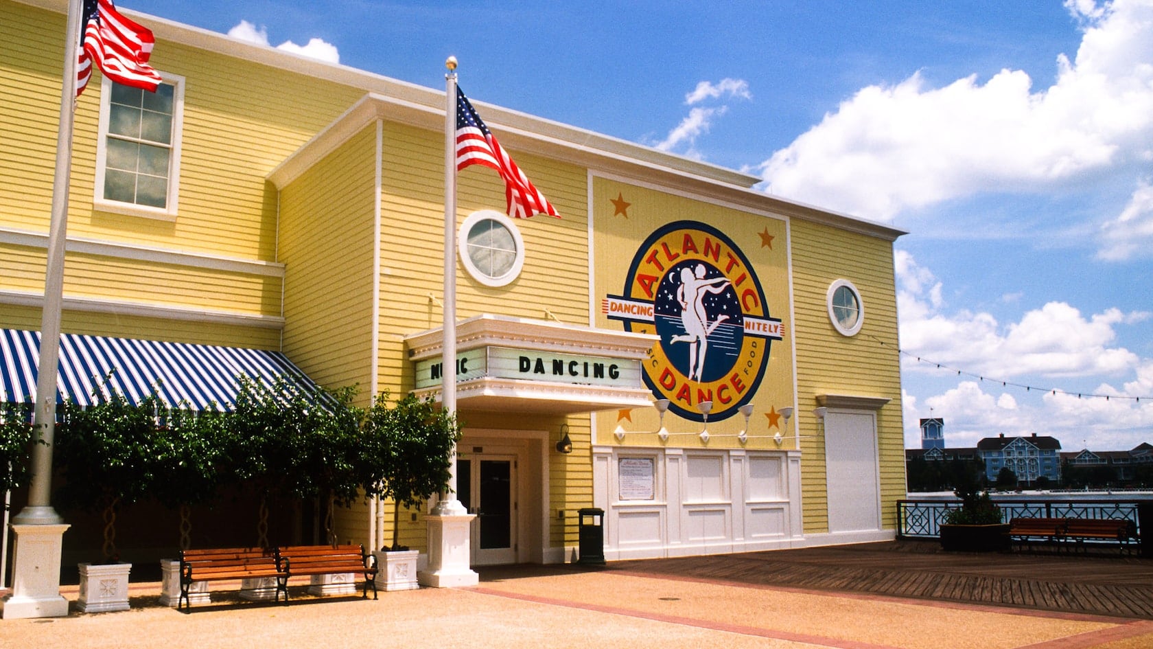 The yellow clapboard entrance of Atlantic Dance Hall on a sunny day, with a marquee reading MUSIC and DANCING, a large round Atlantic Dance logo showing a dancing couple, two American flags, benches and a boardwalk running toward the lake