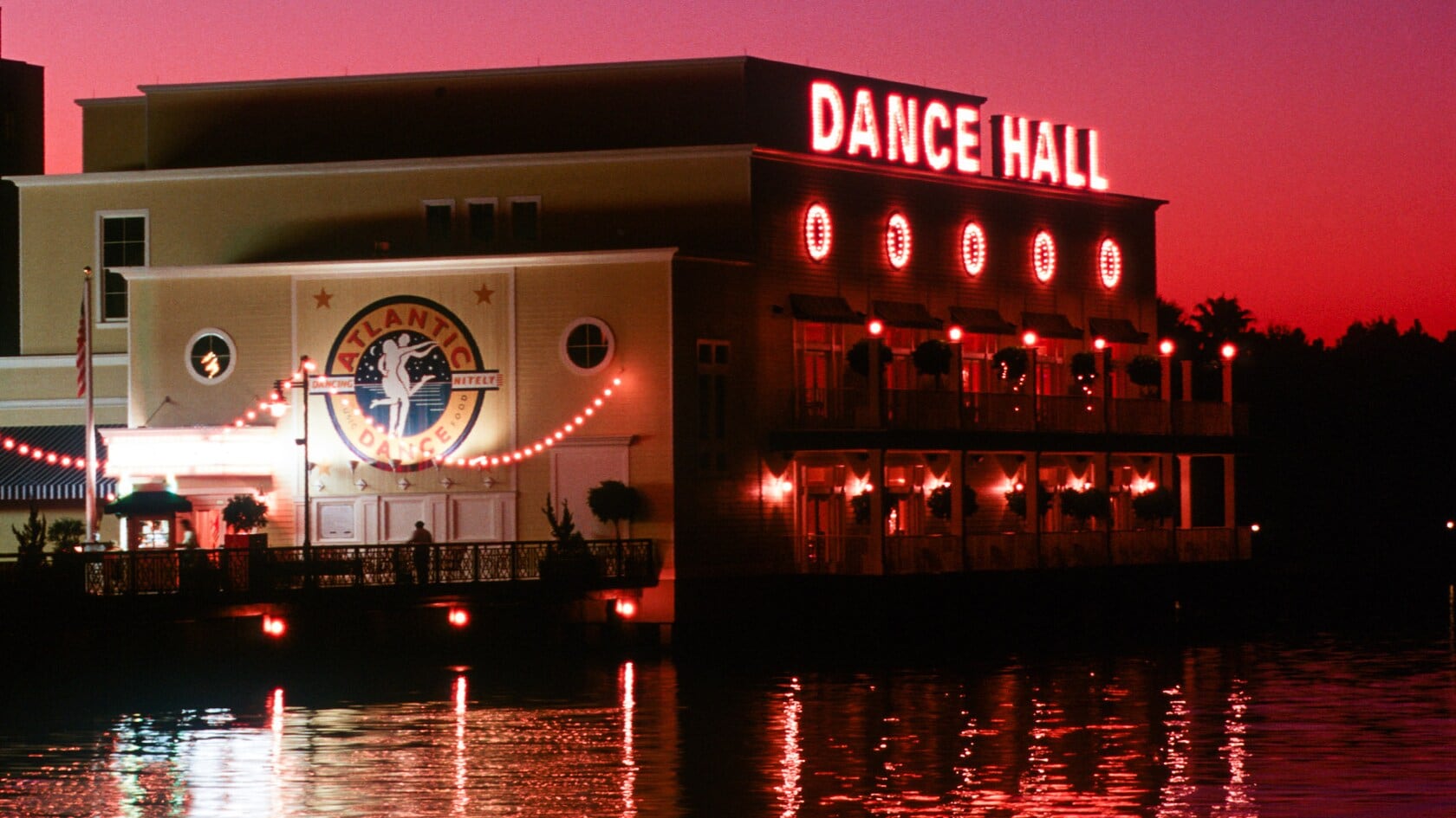 Atlantic Dance Hall at dusk under a pink sky, with red DANCE HALL letters lit on the roof, round lit windows along the waterfront side, and the Atlantic Dance logo on the yellow facade, with the lights reflected in the lake