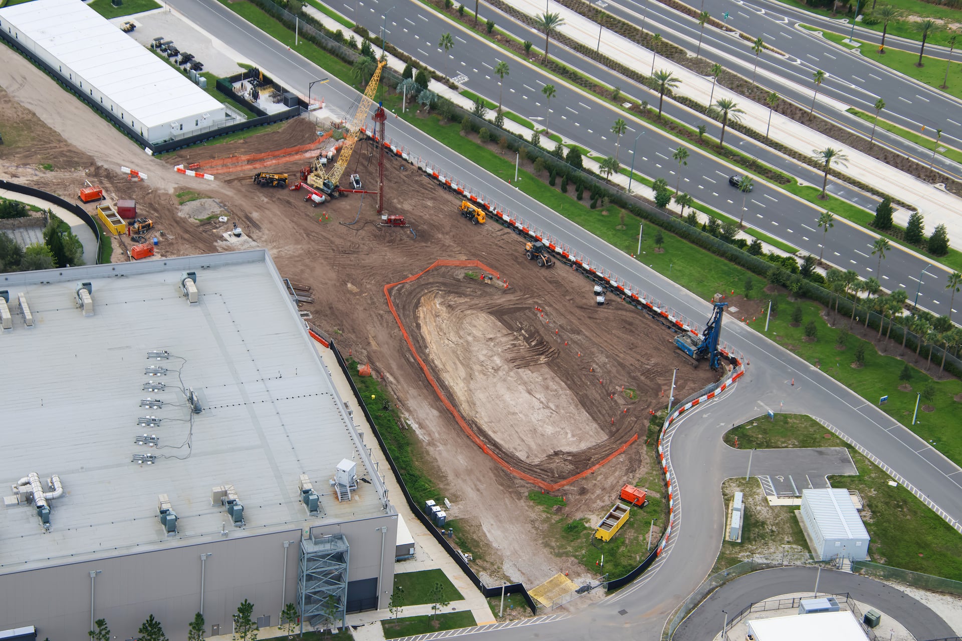 Aerial view of the same construction site looking toward a multi-lane road, with an oval pit fenced in orange, a yellow lattice crane and a red rig at the top of the pad, a blue rig at the corner by the road, and the roof of a large gray building at lower left