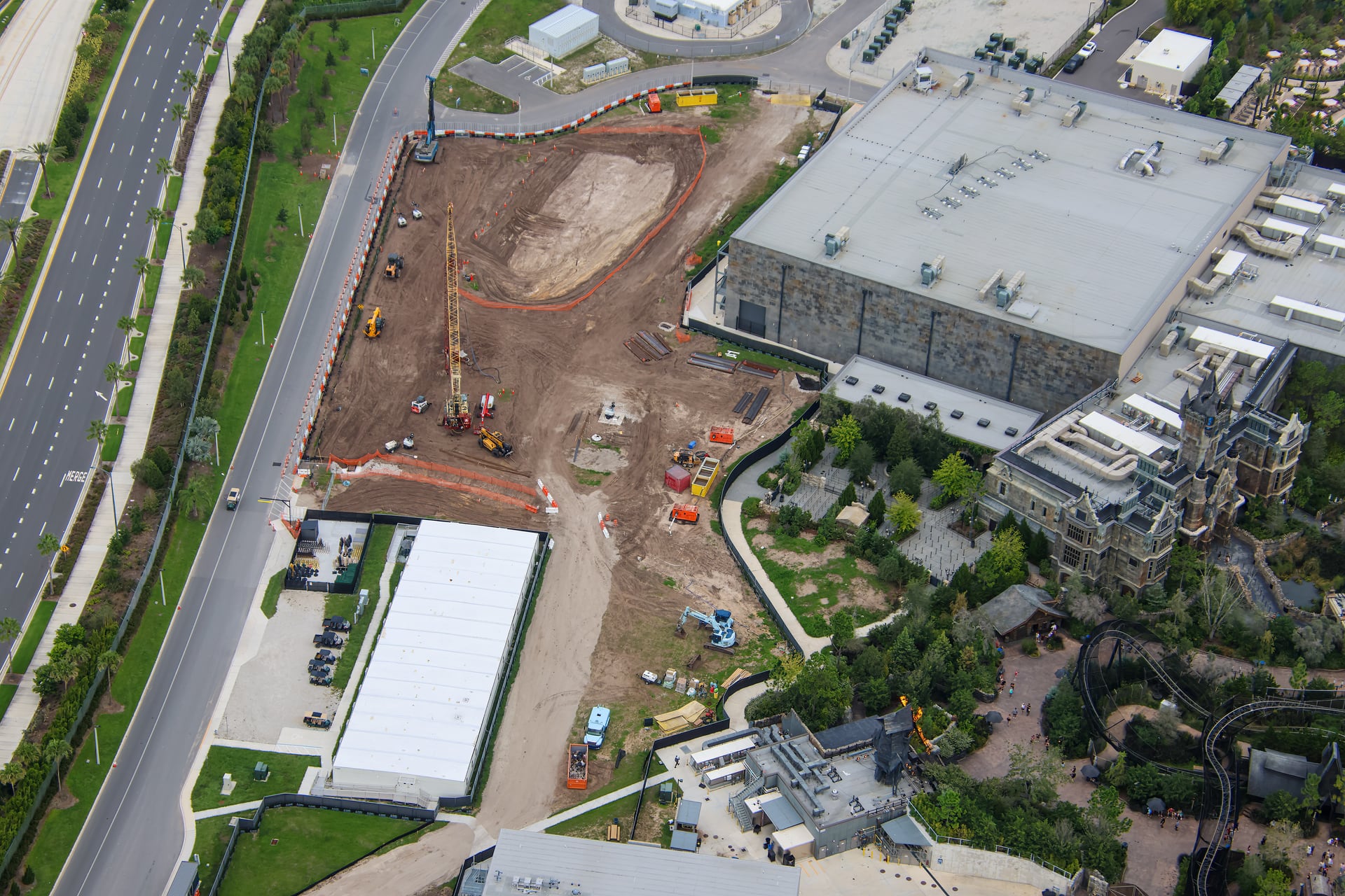 Aerial view of a large dirt construction site beside a gray show building and a Gothic manor, with an oval excavated pit fenced in orange, a tall yellow crane near the middle, a blue rig by the road, white office trailers, and a multi-lane road running along the left