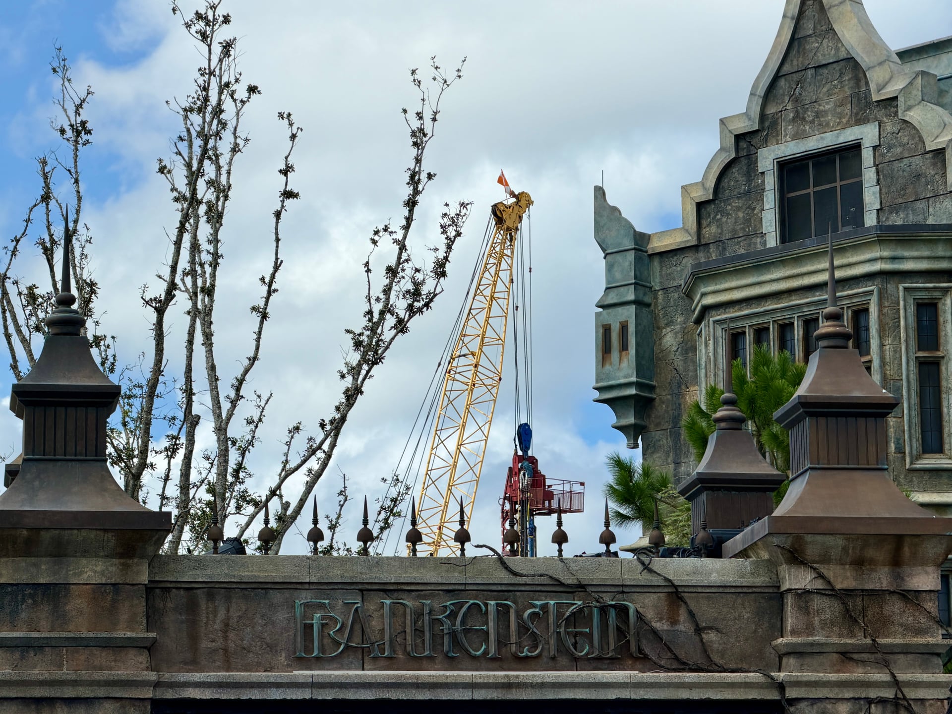 A yellow lattice crane with a red pile driving rig hanging from it, rising above a stone gate lettered with the word Frankenstein, with iron spikes along the top of the wall, bare trees at left and the corner of a Gothic stone manor at right