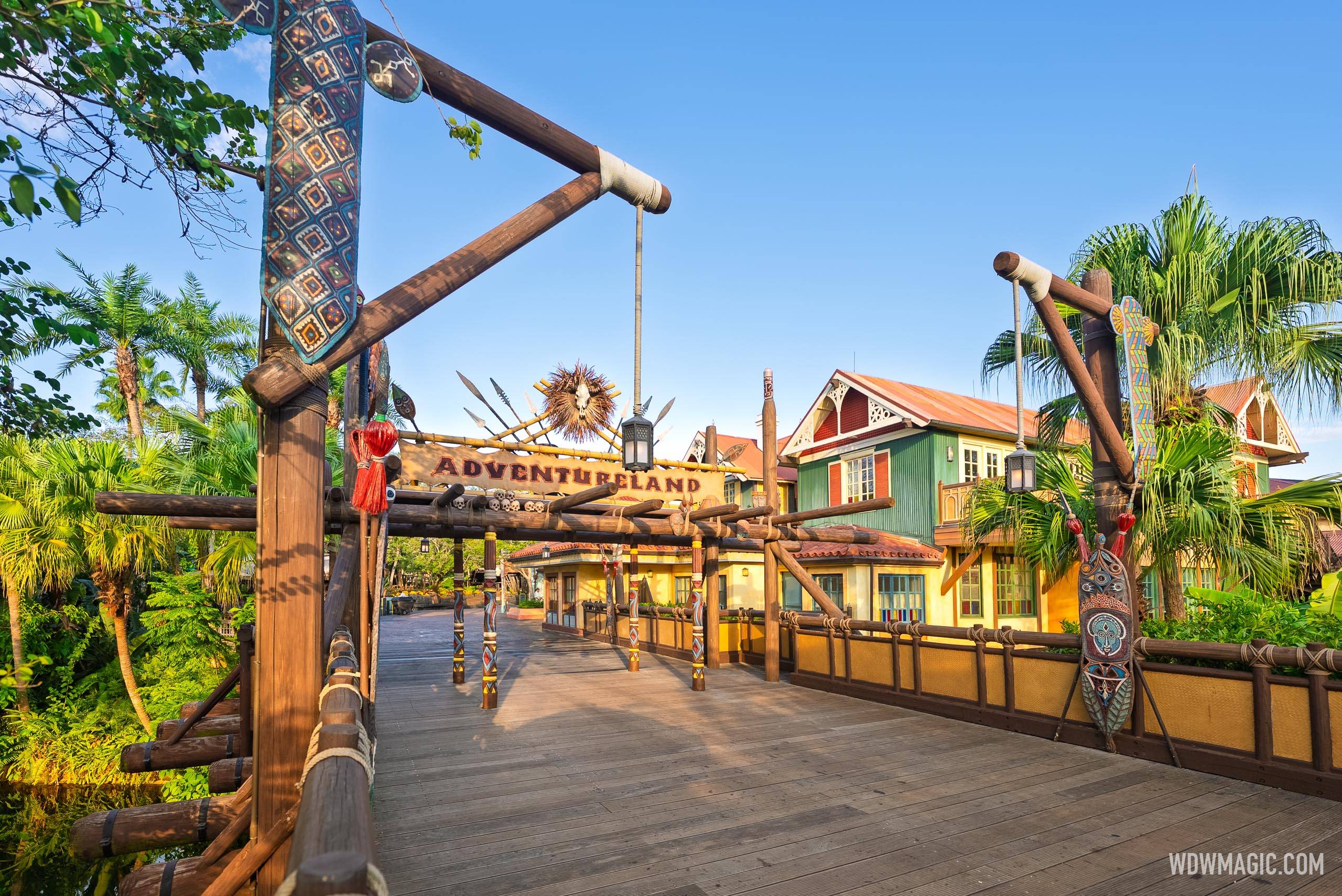 The wooden bridge into Adventureland at Magic Kingdom on a sunny day, with the Adventureland sign and a burst of spears and dried palm fronds over a timber gateway, green and yellow colonial style buildings on the right and palms on both sides