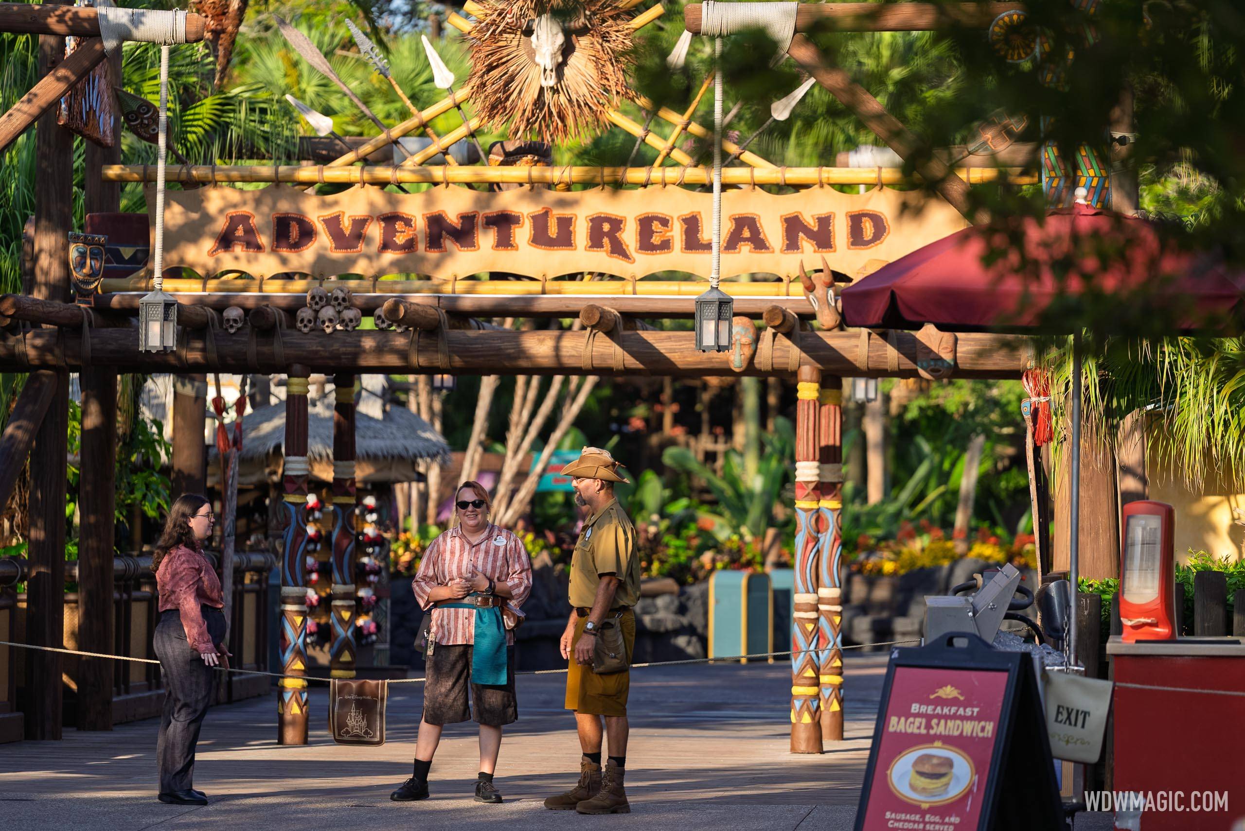 Three cast members in costume chatting under the Adventureland sign, with carved skulls and masks on the timber gateway, a rope strung across the walkway behind them, and a red sign advertising a breakfast bagel sandwich in the right foreground