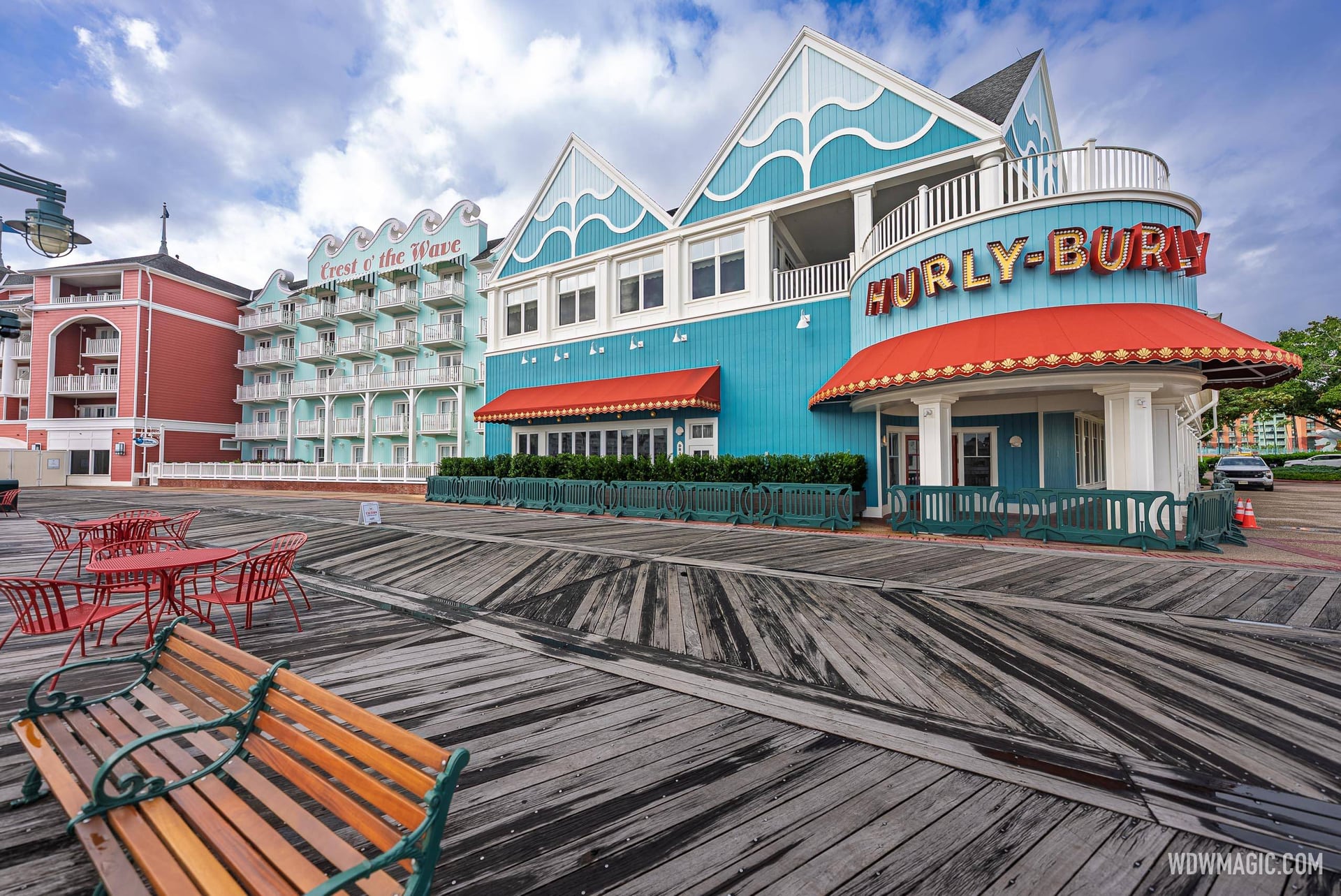 A teal and white Victorian style building on the BoardWalk with a bulb-lined HURLY-BURLY marquee sign and red scalloped awnings, green barricades along its front, wooden boardwalk planks and a bench in the foreground