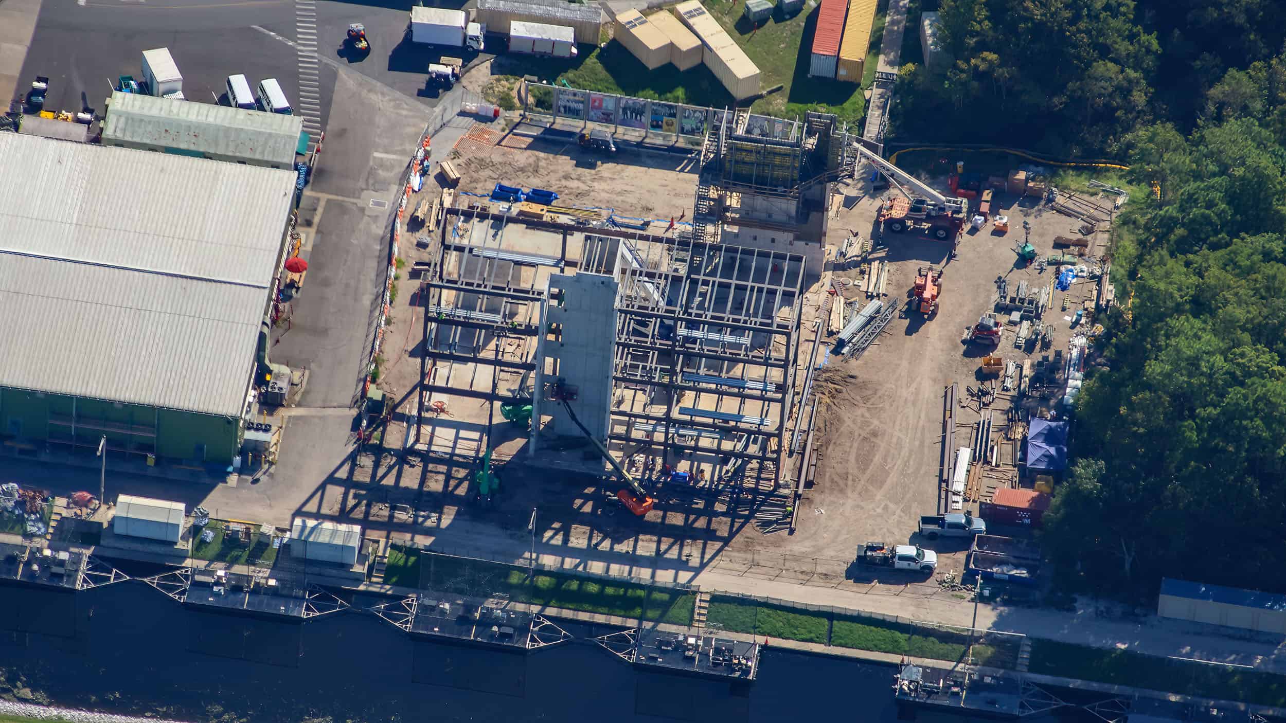 Aerial view looking down on a steel frame several stories tall with a pale precast concrete tower rising at its front edge and an orange boom lift parked at its base, a large grey roofed warehouse with green walls to the left, a dirt yard of stacked materials and pickup trucks to the right, and a row of pontoon barges moored along a canal at the bottom of the frame