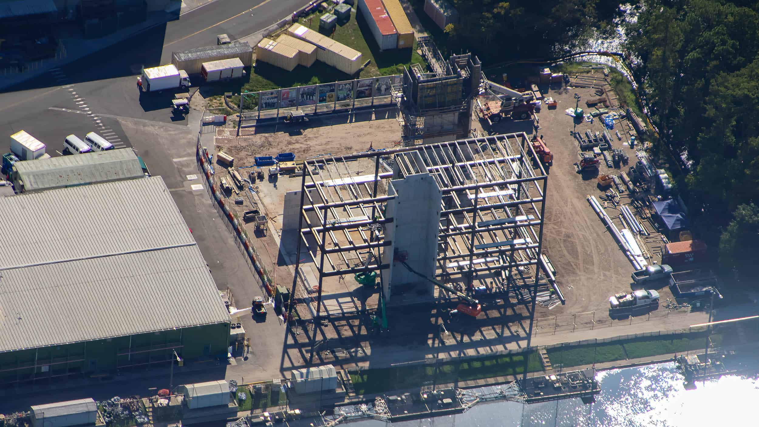 Closer oblique aerial of the same site showing the steel frame and the tall concrete tower rising along its front face, a second scaffolded structure at the back of the lot, a fence hung with large printed panels along the rear edge, the green walled warehouse at left and sunlight glinting off the canal at lower right
