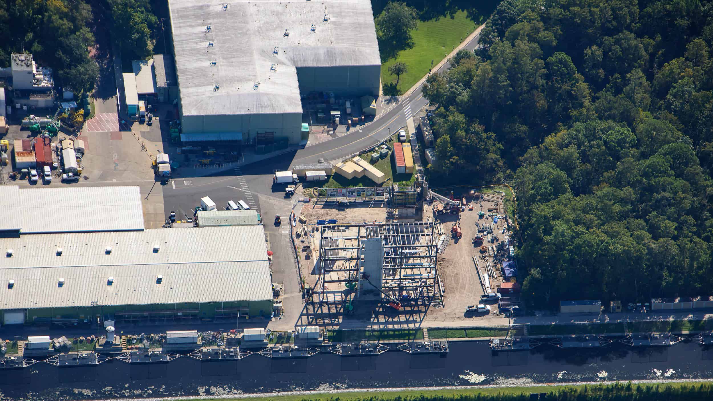 Wide aerial showing the construction site in context: two large grey roofed warehouses at left and top, a backstage road curving past, dense green woods filling the right side, and the steel frame with its concrete tower at center just above a long line of moored barges on the canal