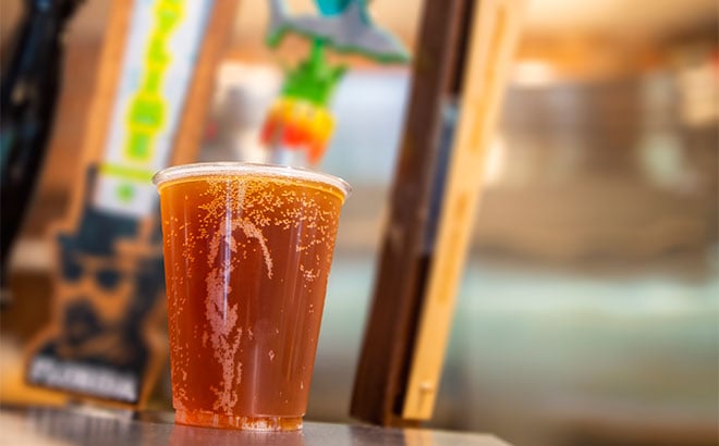 A clear plastic cup of amber beer with foam clinging to the inside, sitting on a bar counter in front of blurred colorful tap handles, one of them reading Florida