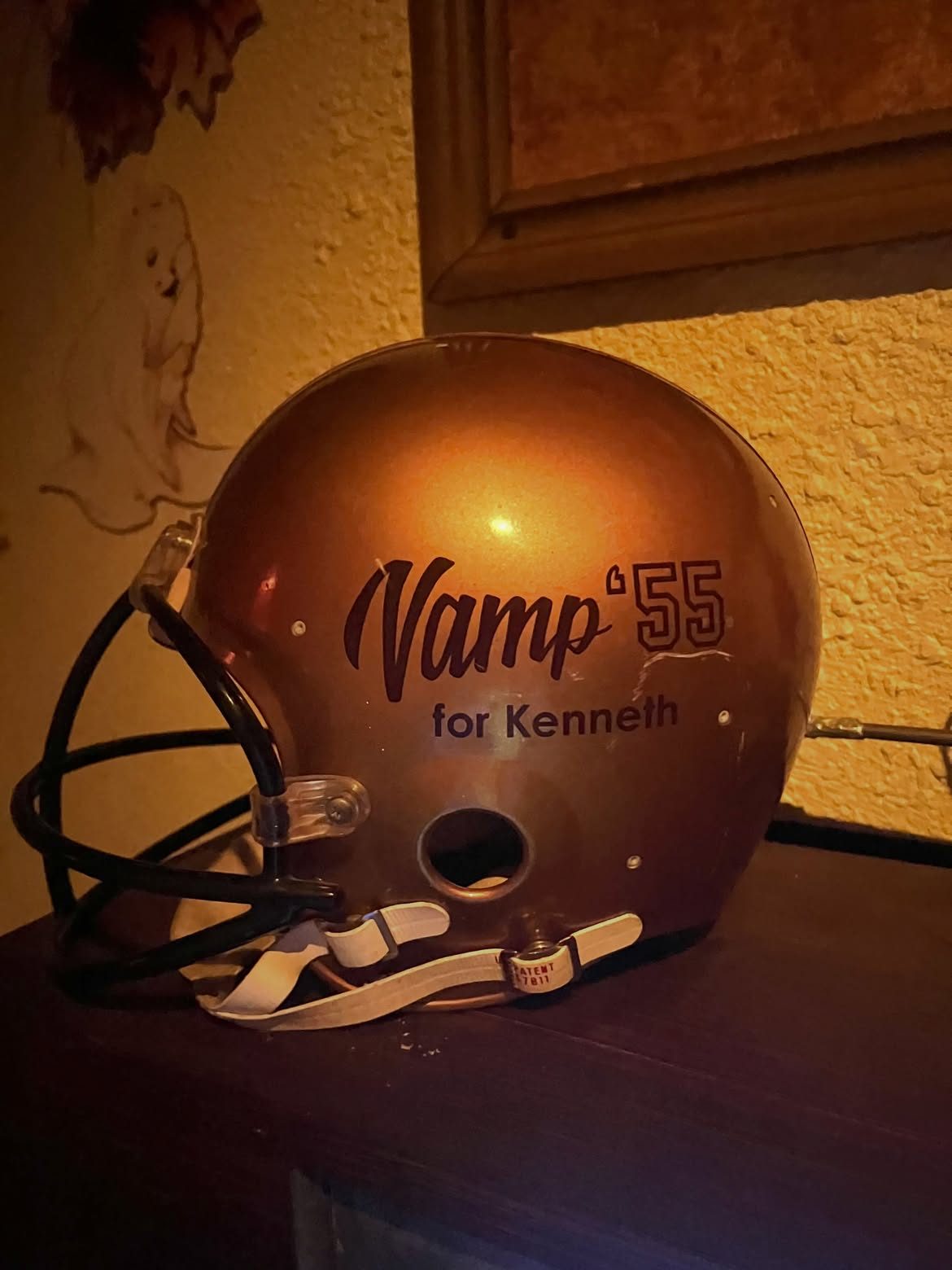 Close-up of a gold football helmet with a black facemask, printed with a script Vamp logo, a block-numeral 55 and the words for Kenneth below it