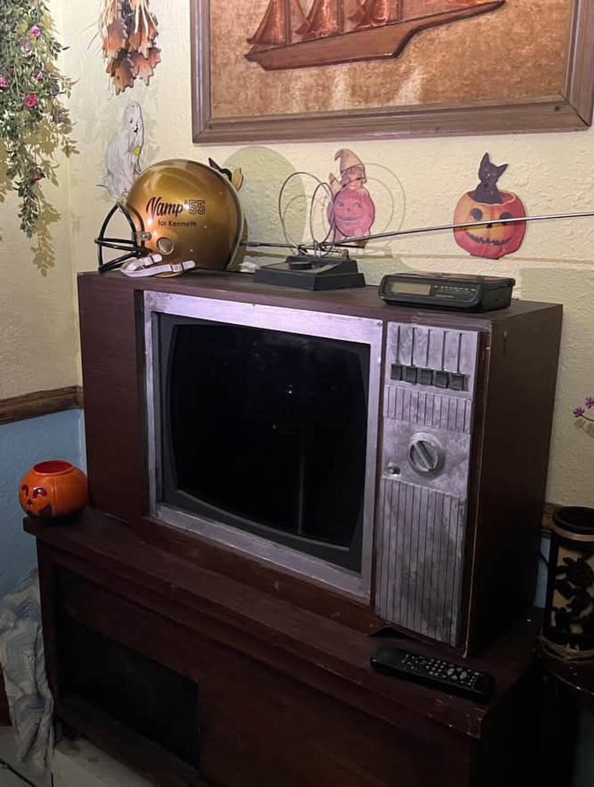 A gold football helmet reading Vamp 55 for Kenneth sits on top of a wood-cabinet console TV with rabbit ear antennas, a clock radio and vintage Halloween cutouts on the wall behind it