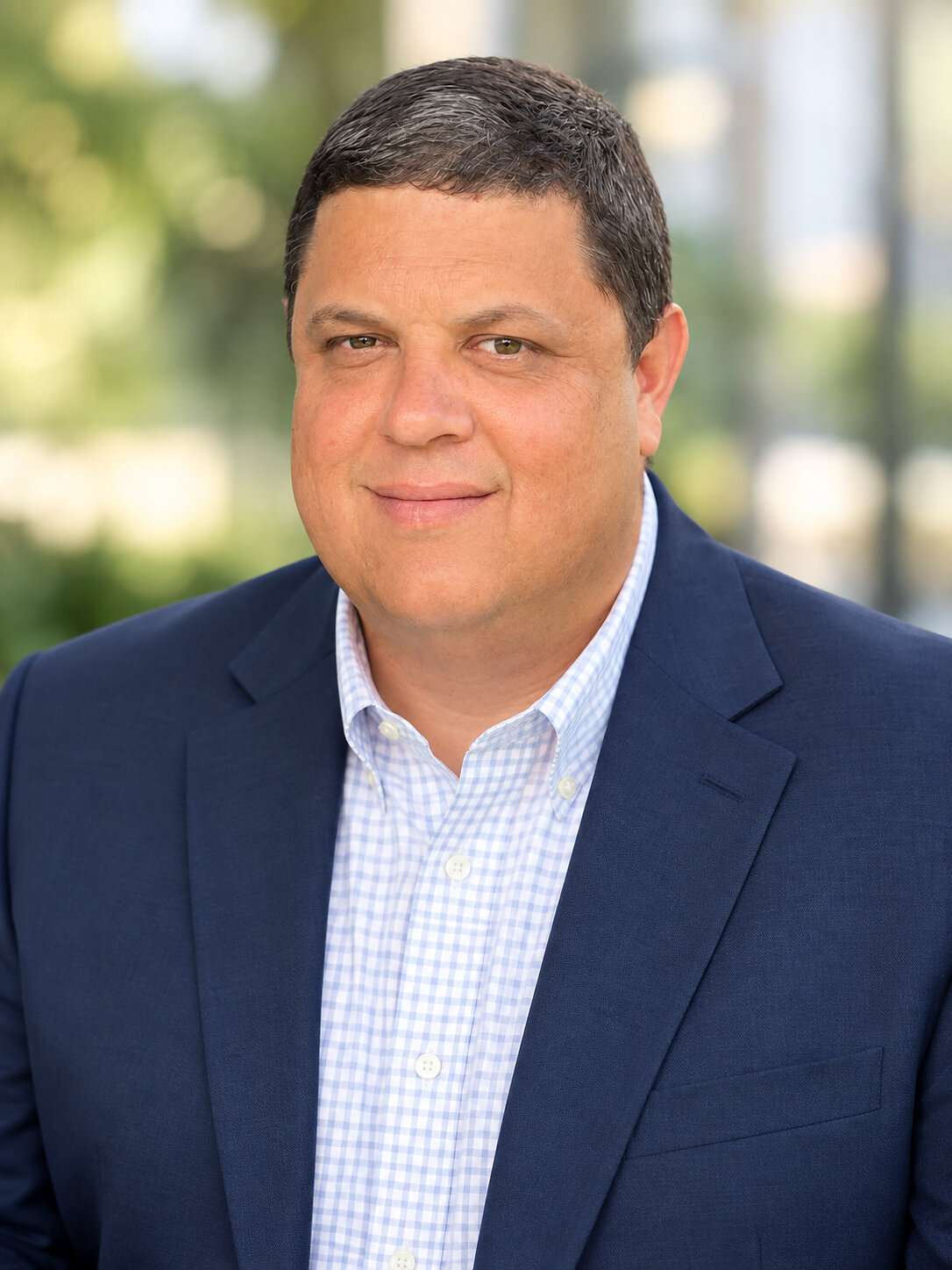Corporate headshot of Kyle Miller, chest up, short dark hair and a slight smile, wearing a navy blazer over a light blue and white checked open collar shirt, photographed outdoors against a blurred background of greenery and a pale building facade
