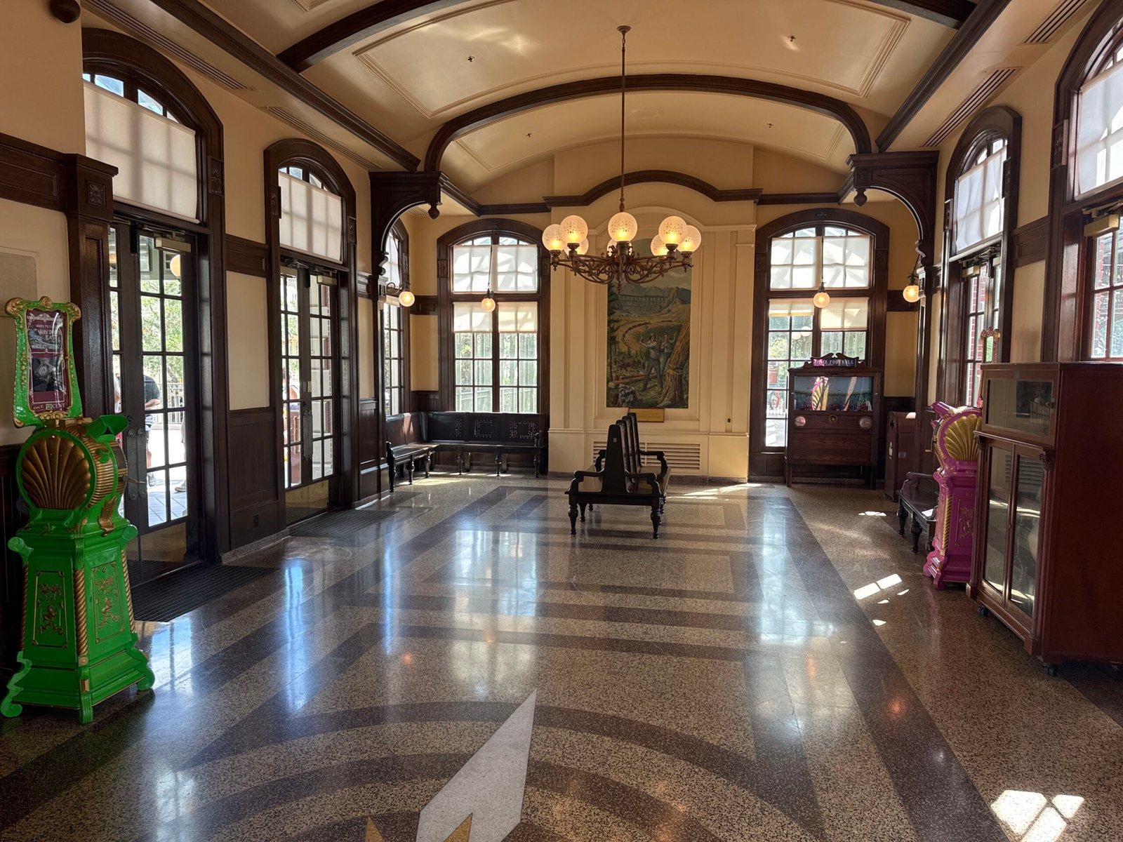 The interior of the Main Street train station at Magic Kingdom, with a polished terrazzo floor, arched windows, a chandelier, wooden benches and a mural on the back wall, a green and gold Mutoscope at left and a pink and gold Mutoscope beside a wooden display cabinet at right