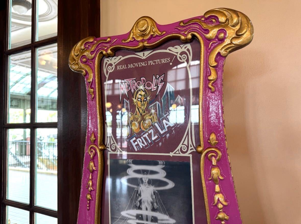The top of a pink and gold Mutoscope with ornate gold scrollwork around a sign reading Real Moving Pictures, Metropolis and Fritz Lang, with an illustration of the film's robot, and a black and white still of the robot surrounded by rings of light below it