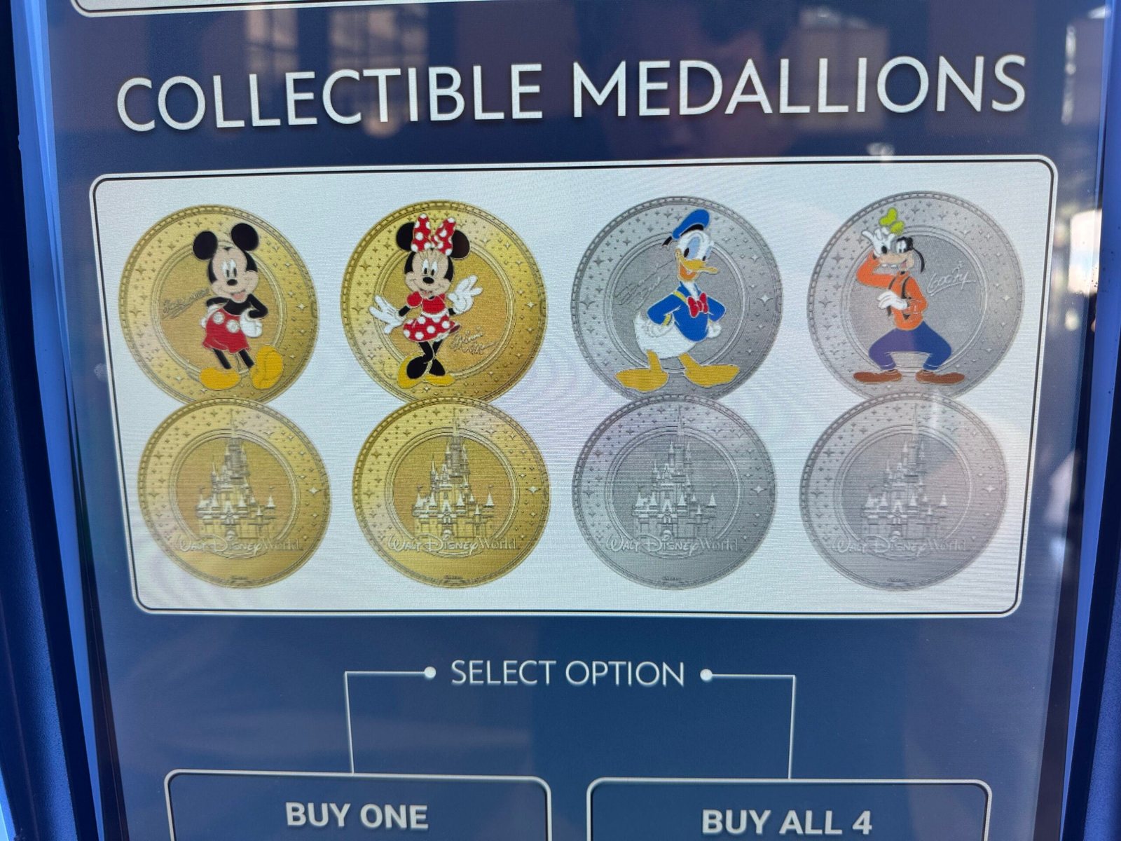 A screen headed Collectible Medallions showing the fronts and backs of four coins: Mickey and Minnie on gold coins and Donald and Goofy on silver coins, each backed by an image of Cinderella Castle, with Select Option, Buy One and Buy All 4 buttons below