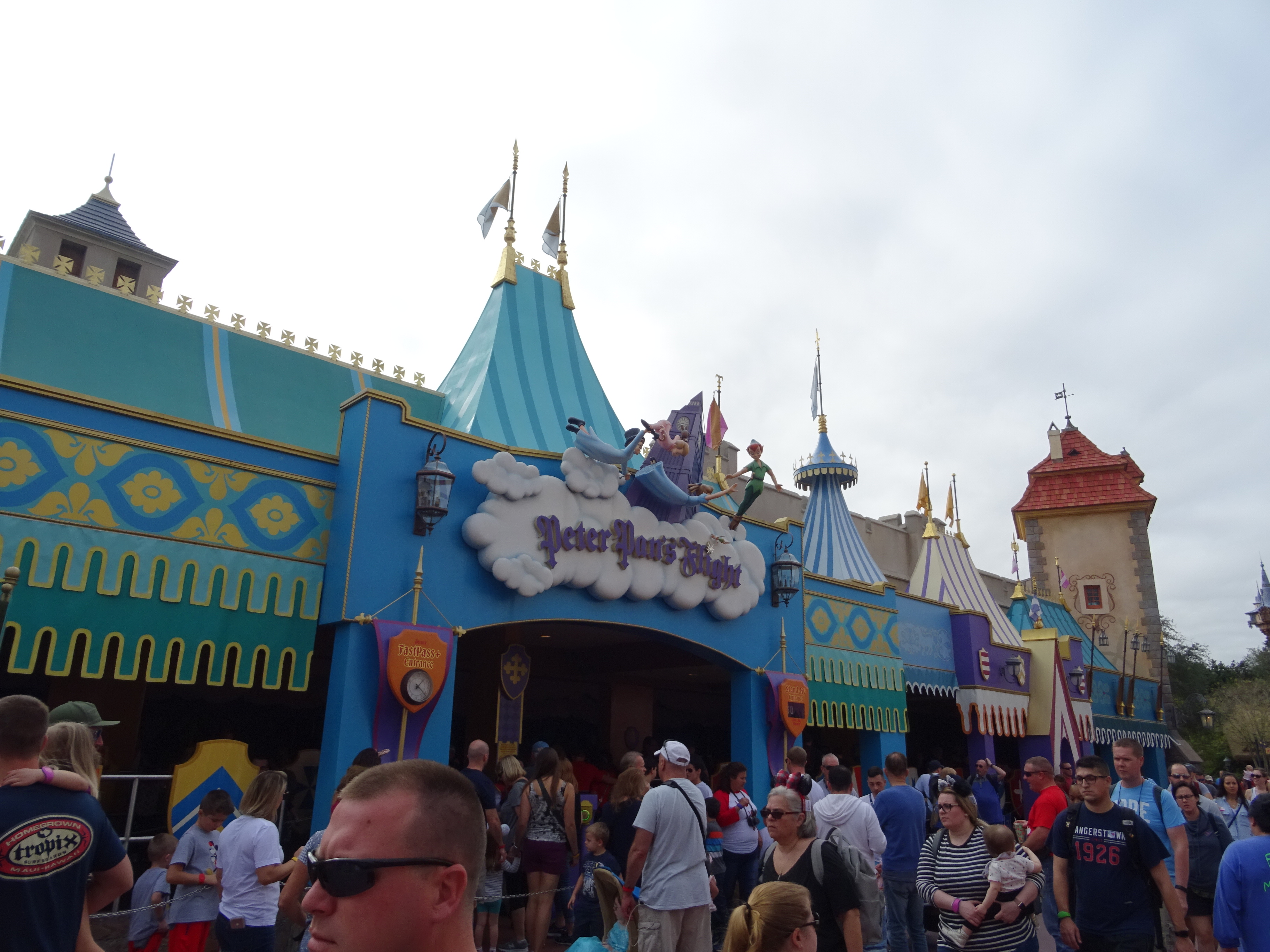 A crowd of guests outside the blue and teal Peter Pan's Flight entrance in Fantasyland, with the cloud marquee above the archway and the striped rooflines of neighboring Fantasyland buildings to the right