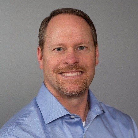 Square corporate headshot of Scott Maupin, chest up and smiling, with short brown hair and a short beard and moustache, wearing a light blue button down shirt against a plain grey studio backdrop