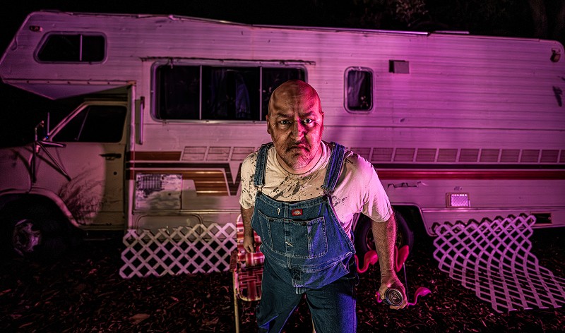 A scowling bald man in stained denim overalls and a dirty white T-shirt stands in front of an old RV lit in pink light at night, holding a can