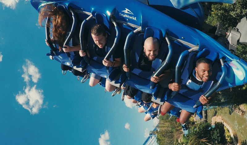 Four riders, three of them smiling, hang face down in a blue flying coaster train marked with the SeaWorld logo, against a blue sky with a few white clouds