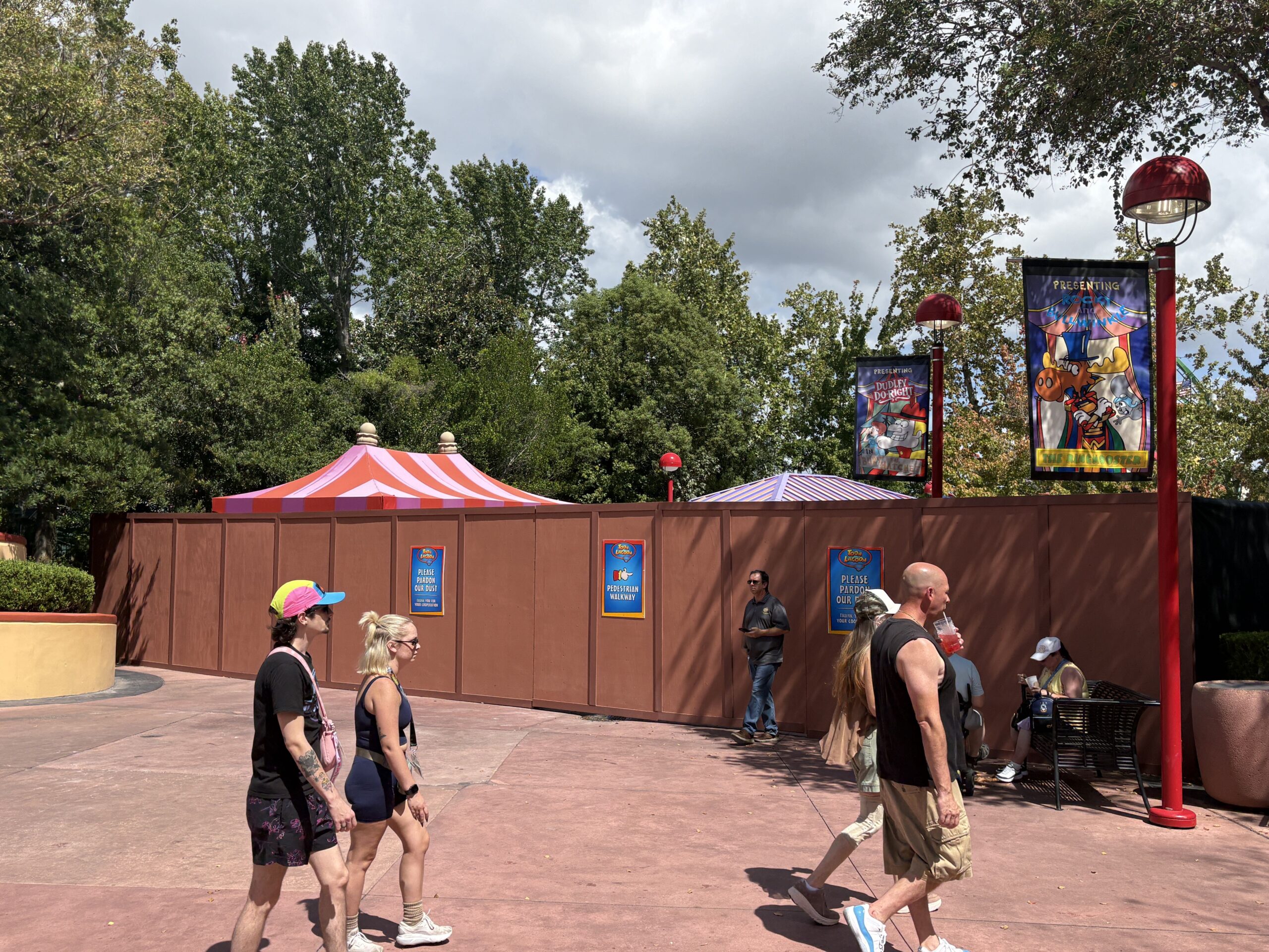 A long brown construction wall running across a Toon Lagoon walkway with guests walking past in the foreground, striped carnival canopies visible behind the wall and lamppost banners for Dudley Do-Right and Rocky and Bullwinkle overhead