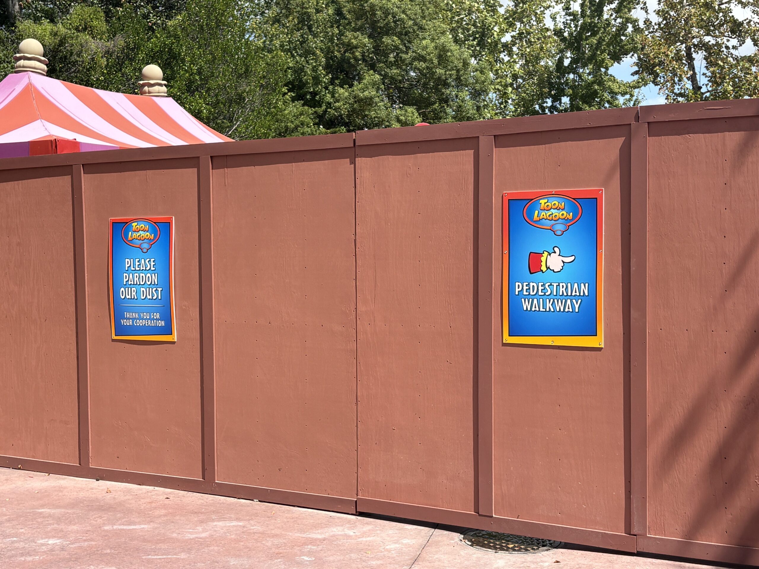 Close view of the brown construction wall with two blue and orange Toon Lagoon signs mounted on it, one reading Please Pardon Our Dust and Thank You For Your Cooperation, the other reading Pedestrian Walkway