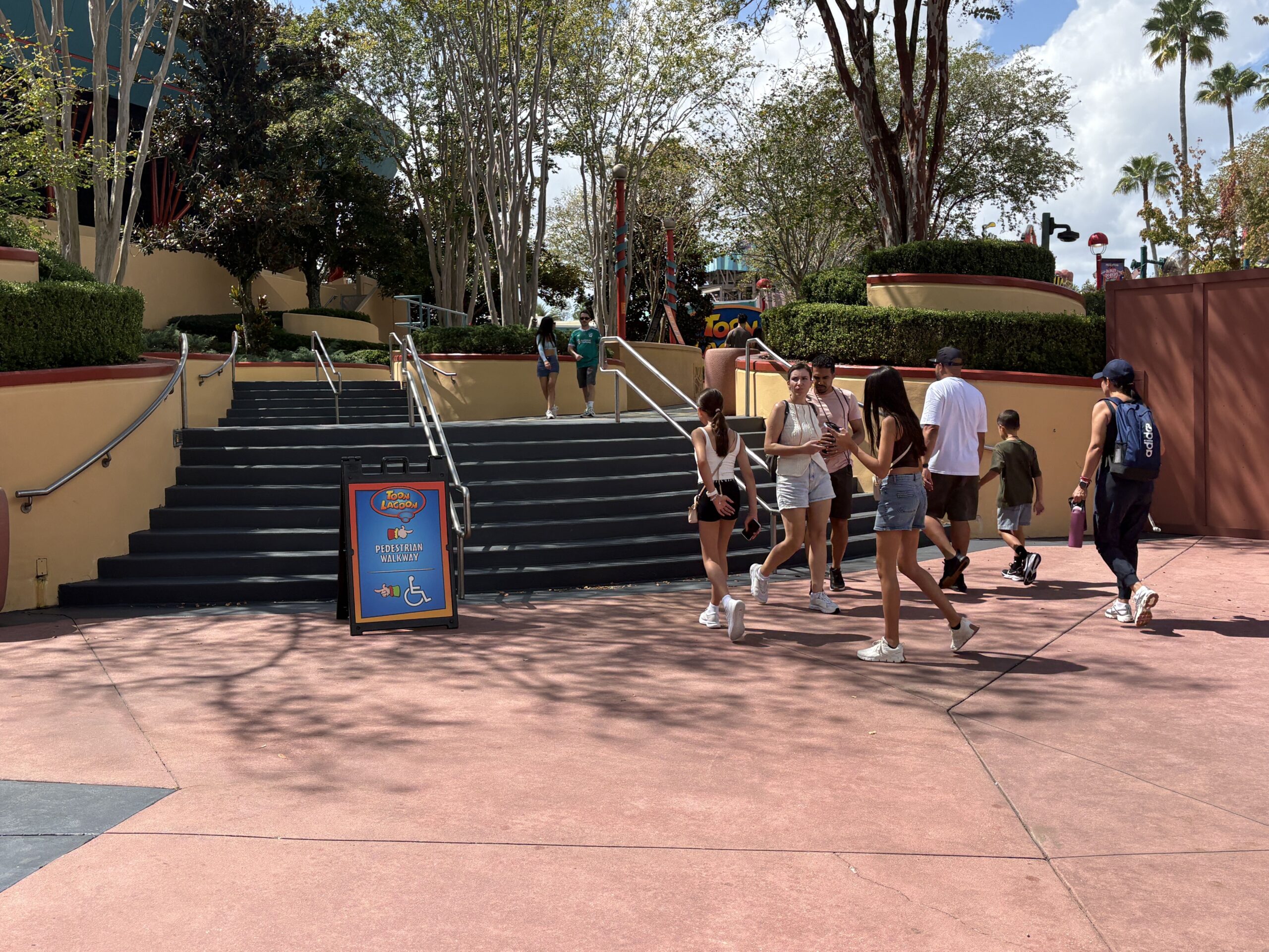 A Toon Lagoon branded A-frame sandwich board reading Pedestrian Walkway with a pointing glove and a wheelchair symbol, standing at the base of a wide staircase as guests walk past