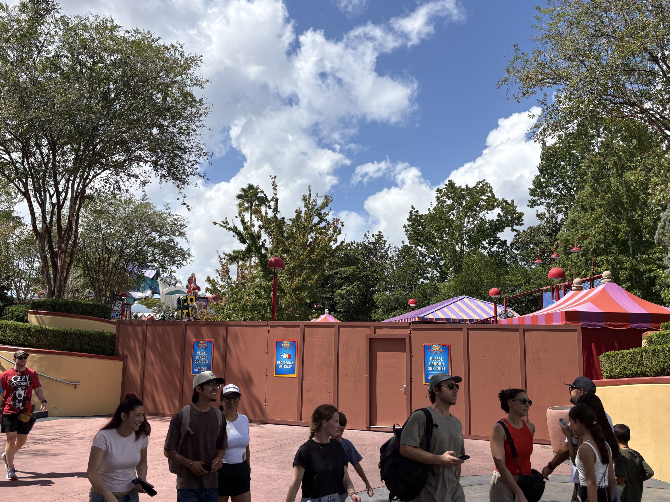 Wide view of the construction wall with an access door set into it, three striped carnival game canopies in pink, orange and purple rising behind, and guests walking along the pink pavement in front