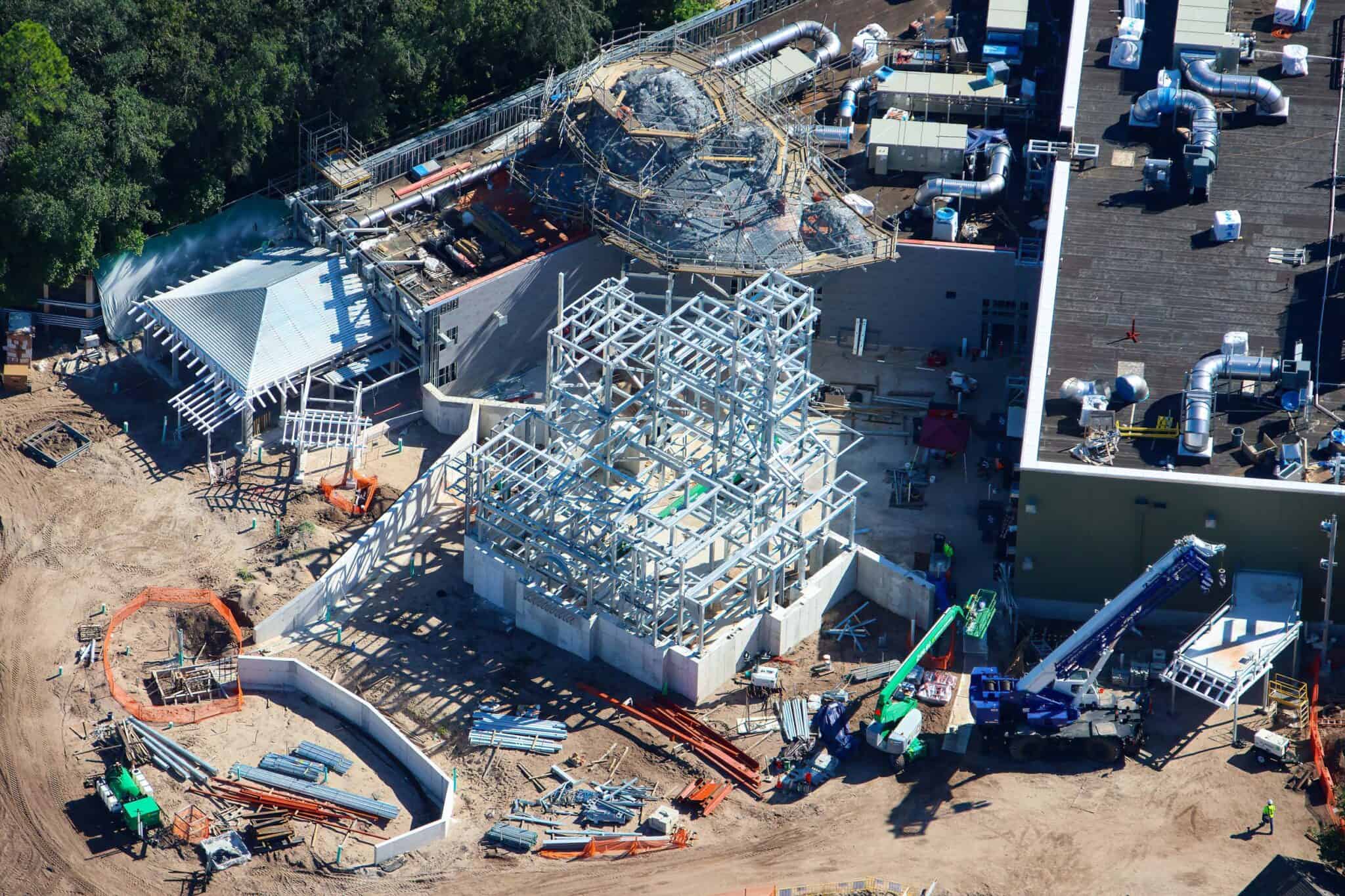 Aerial view of a bare silver steel frame standing on a poured concrete base, shaped like a multi level house with a tower at one end, in front of a flat roofed show building covered in ductwork and HVAC units, with a scaffolded rockwork mound behind it and a blue crane and stacked steel on the dirt nearby