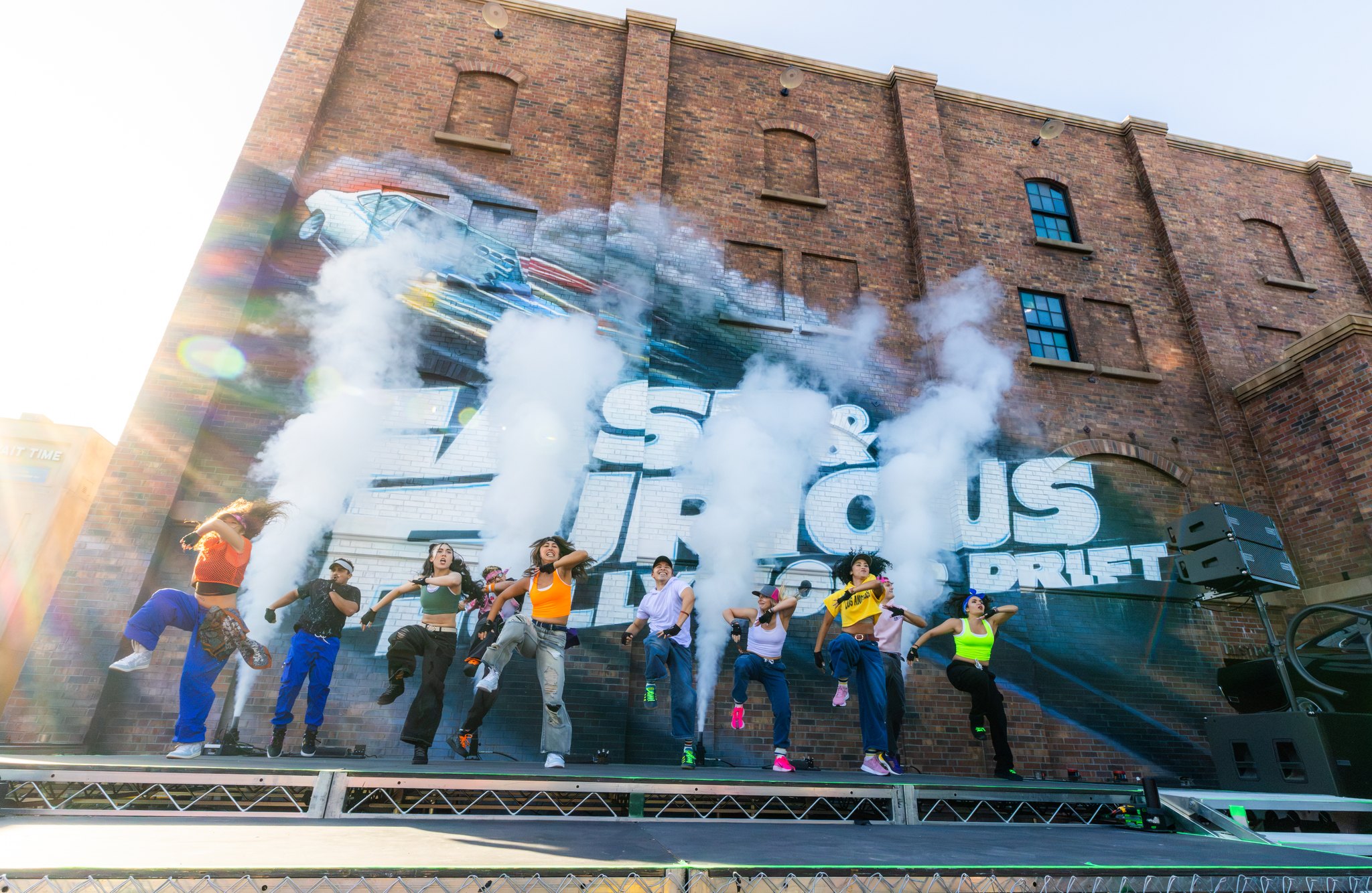 A line of dancers in bright streetwear leap and pose on an outdoor stage in front of a brick wall painted with a large Fast and Furious Hollywood Drift mural, with thick white smoke drifting across the letters and sun flaring at the top left