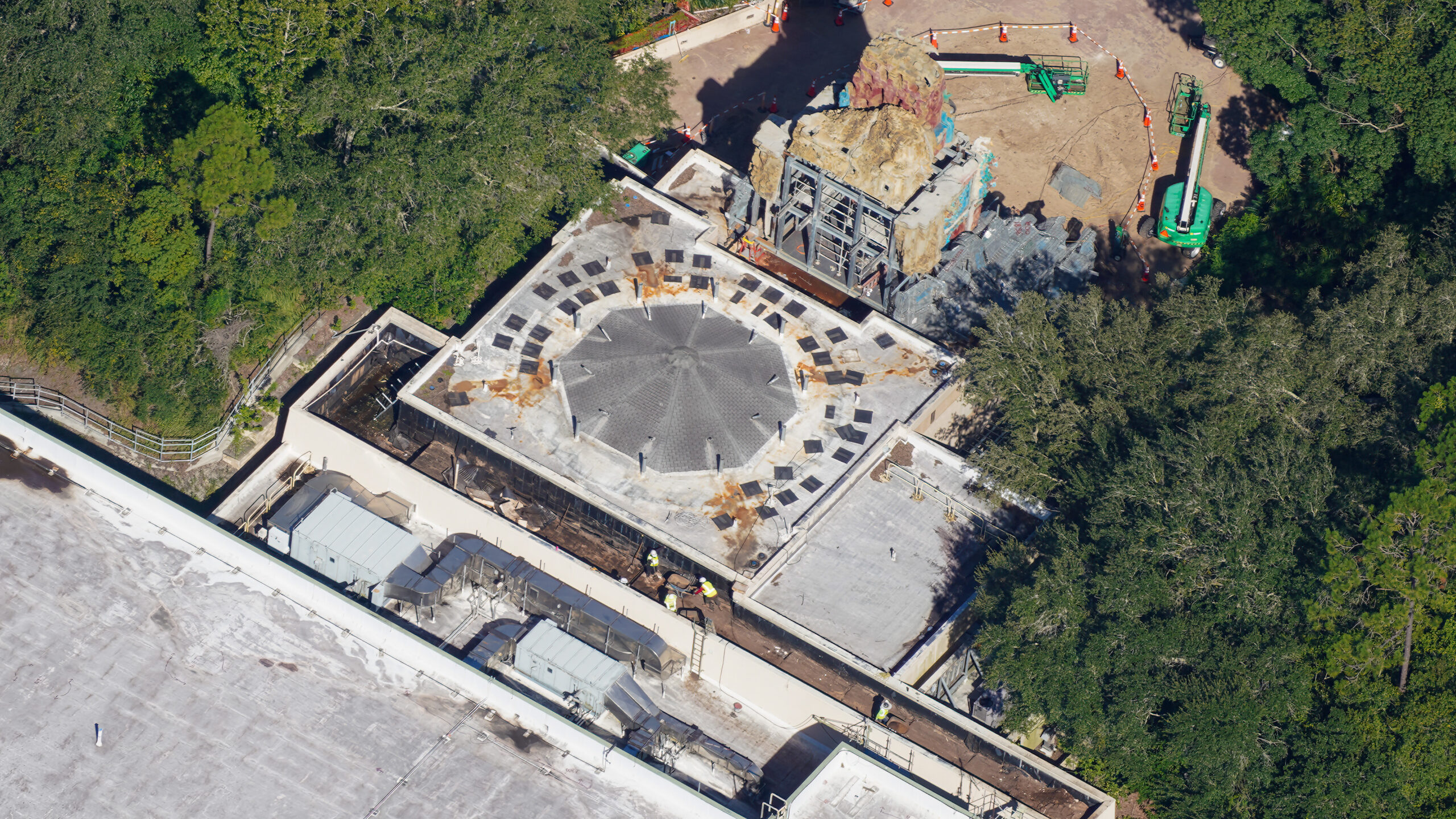 Aerial view of a large flat roof with an octagonal paved plaza at its center ringed by dark panels and short posts, bare dirt and rust stained runoff around the edges, three workers in yellow vests in a trench at the lower edge, and part of the carved temple facade with green boom lifts along the top edge