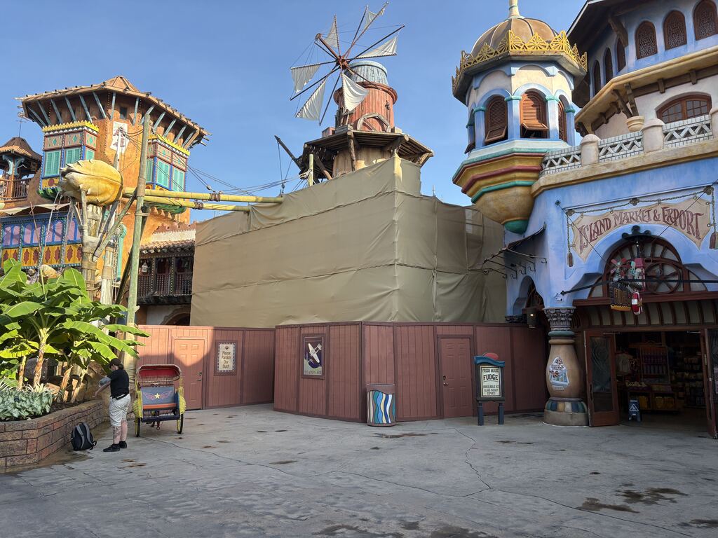 Beige scrim over a windmill topped structure behind brown construction walls, with the Island Market and Export storefront open and stocked at right and a Please Pardon Our Dust sign on the wall