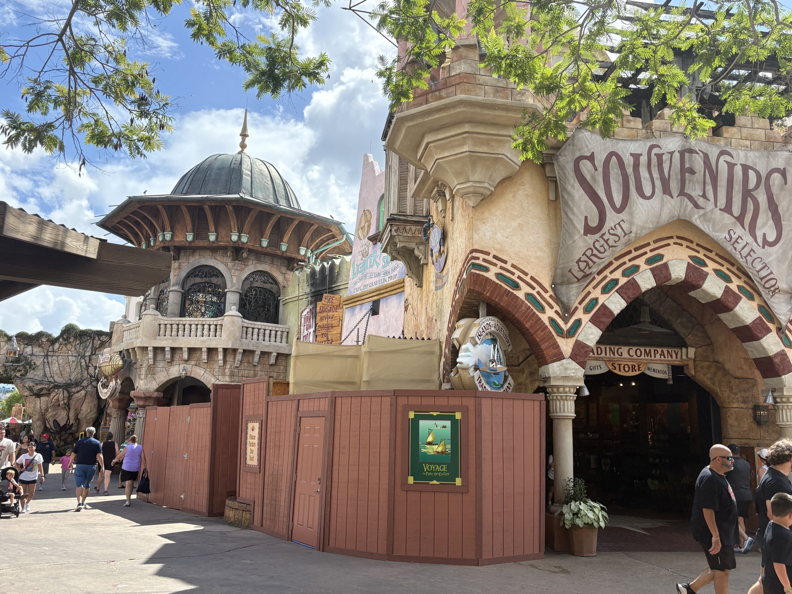 Brown construction walls curving around the left side of the Islands of Adventure Trading Company, with the Souvenirs Largest Selection awning above the open store arch and guests walking past
