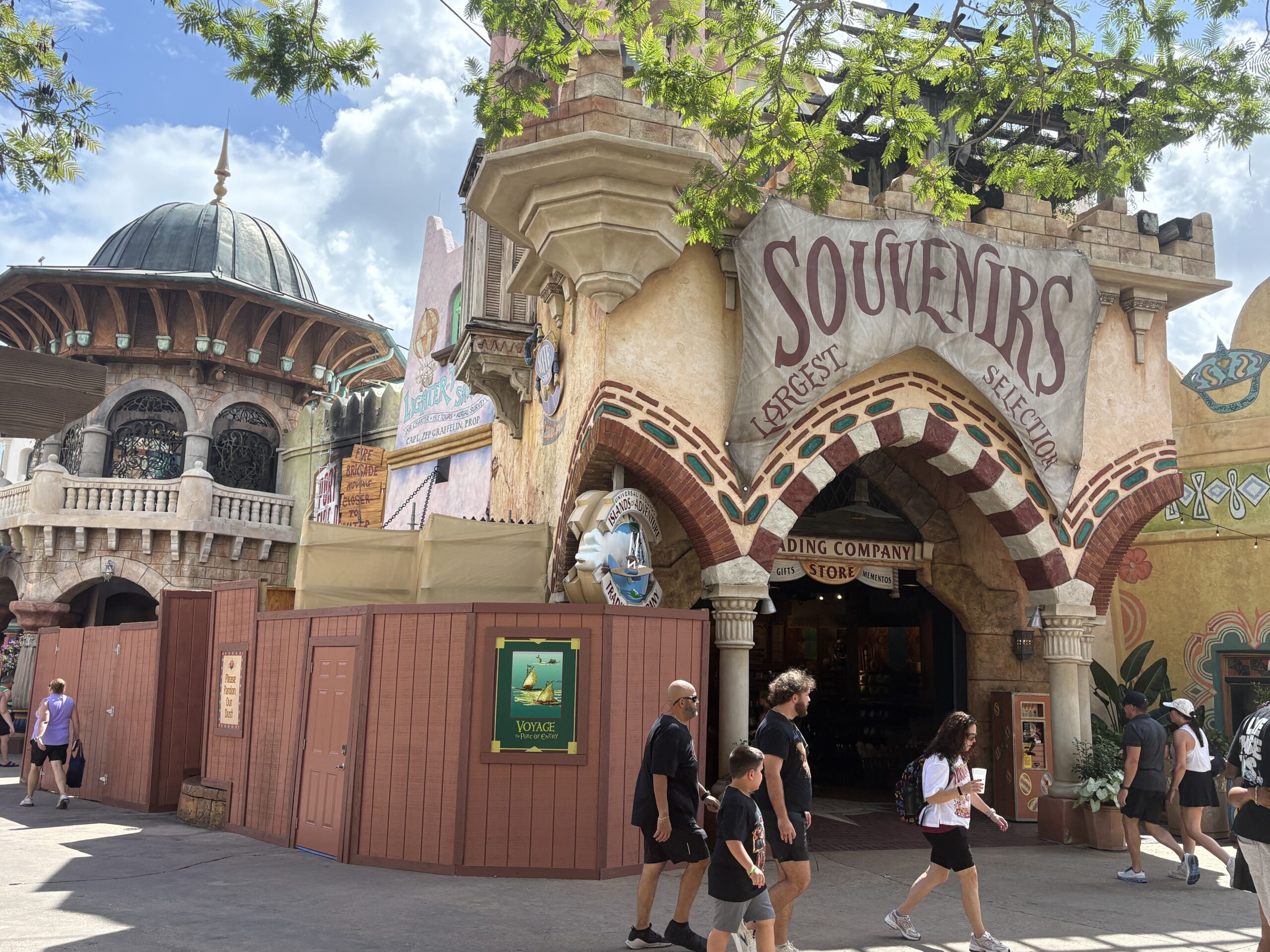 Wider view of the Islands of Adventure Trading Company entrance arch with guests walking past the open doorway, construction walls at left carrying a Voyage to Port of Entry poster and a Please Pardon Our Dust sign