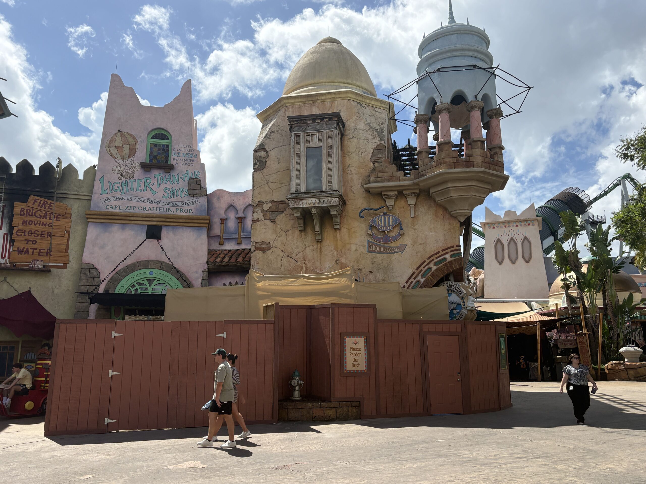 Brown construction walls and beige scrim covering the lower facades in Port of Entry, with the Lighter-Than-Air Ships sign at left and a Kite Shop sign pointing around the corner above the scrim, a framed Please Pardon Our Dust sign on the walls and guests walking past