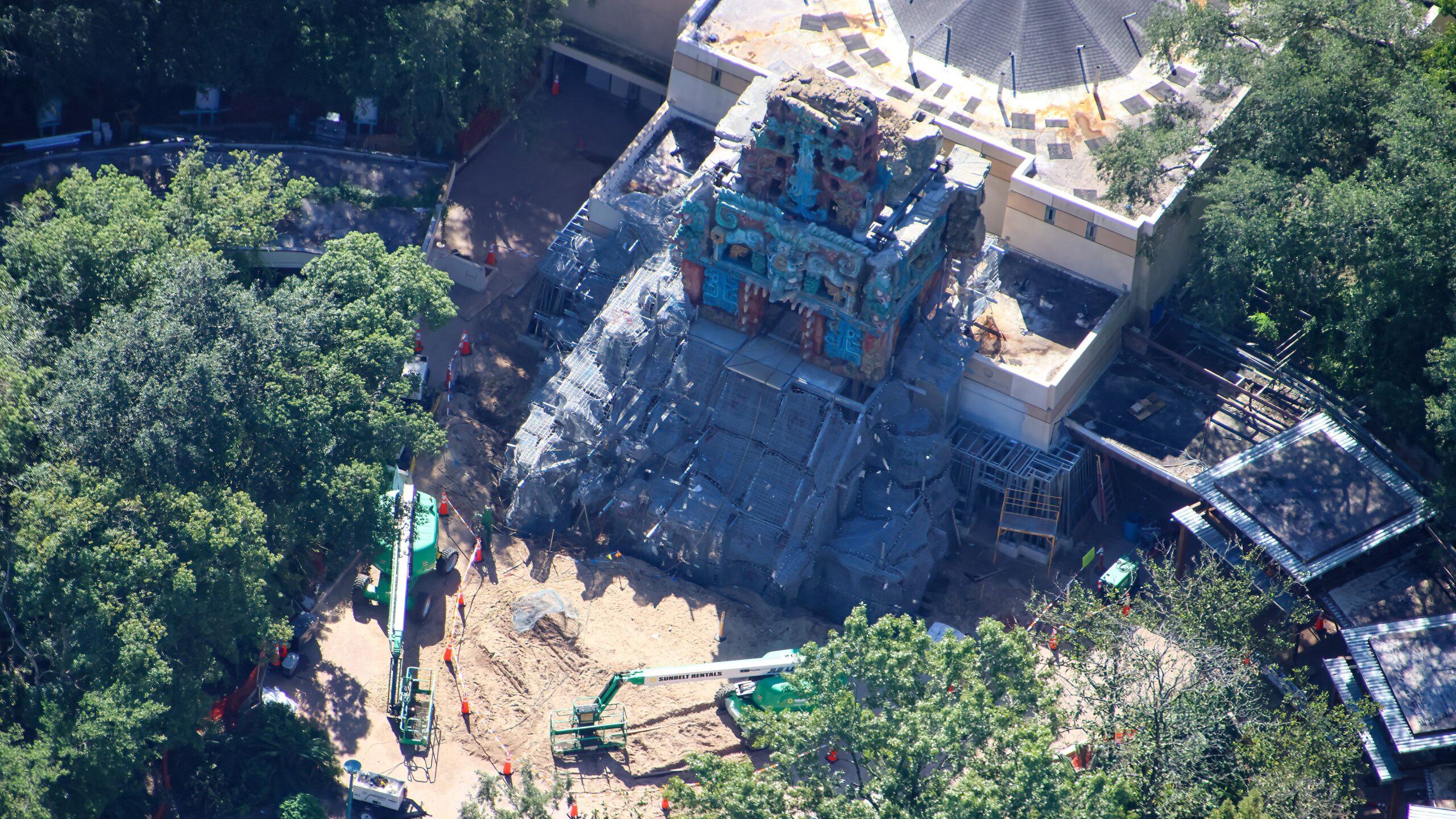 Aerial view of the Maya temple facade on the Indiana Jones ride building, with carved and painted turquoise, red and gold stonework running from the crest down through the middle band, gray unpainted rockwork and wire mesh sloping away below it, and green boom lifts parked on the dirt in front
