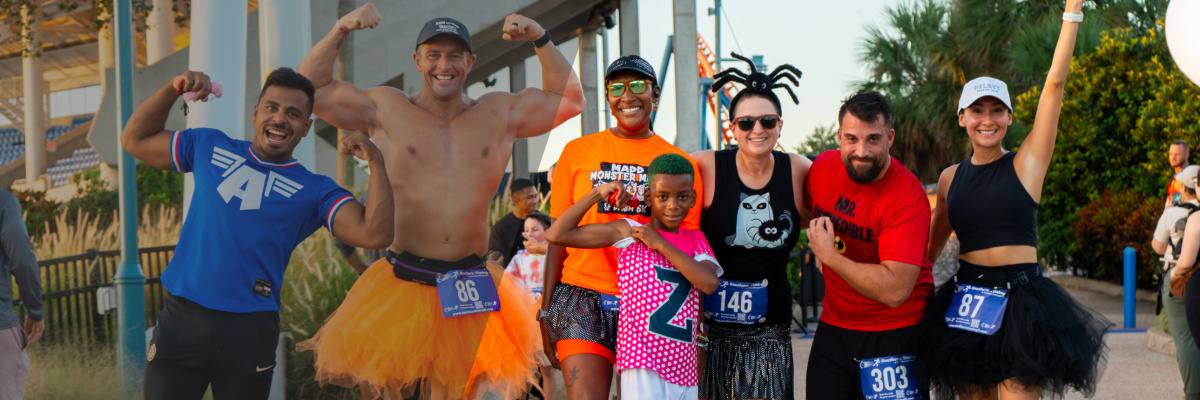 A group of seven adults and children posing together, several wearing race bibs, in an orange tutu, a spider headband, a Captain America jersey and a MADD Monster Mash and Dash race shirt, with roller coaster track and stadium seating behind them