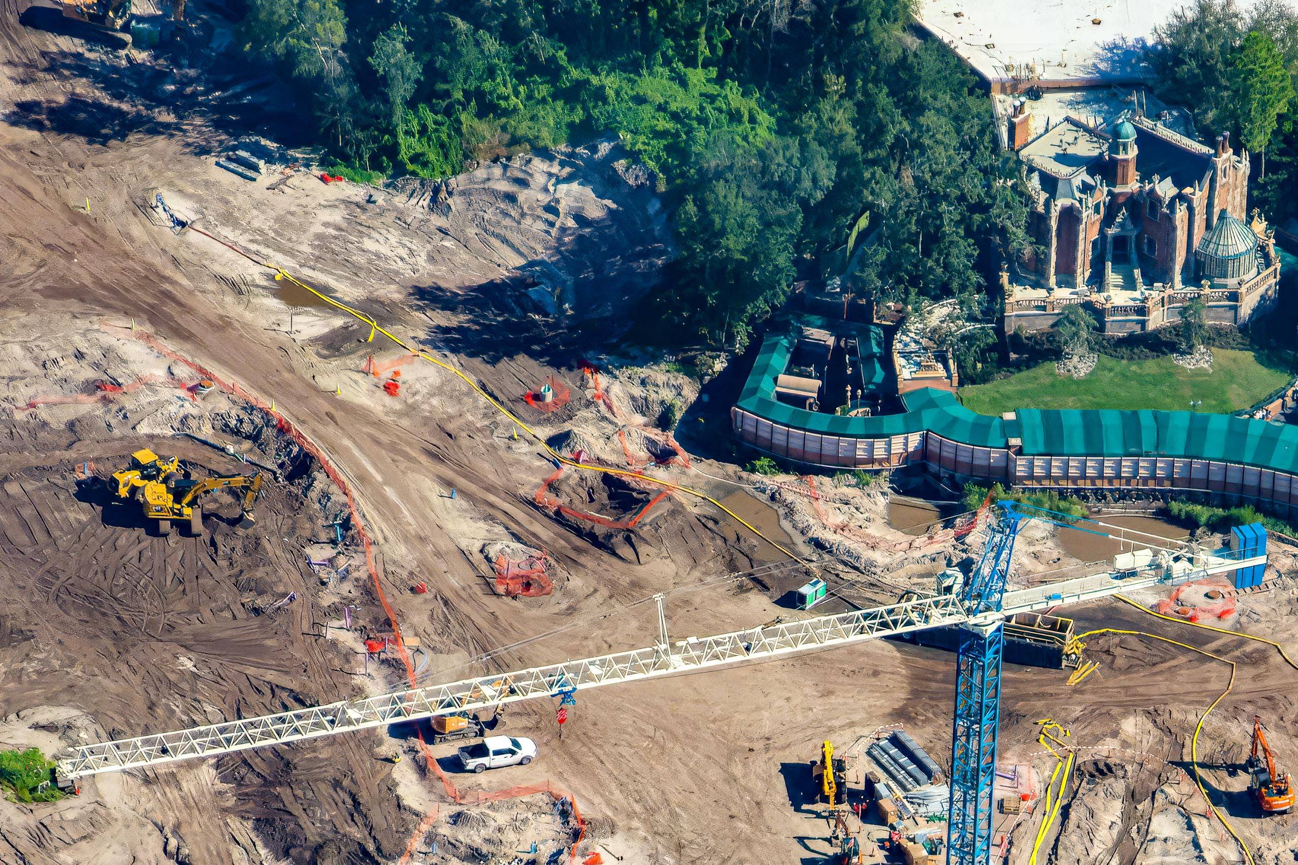 Aerial view of graded dirt, excavators and a blue tower crane on the Piston Peak construction site, with the Haunted Mansion and the green construction walls around the work site at upper right