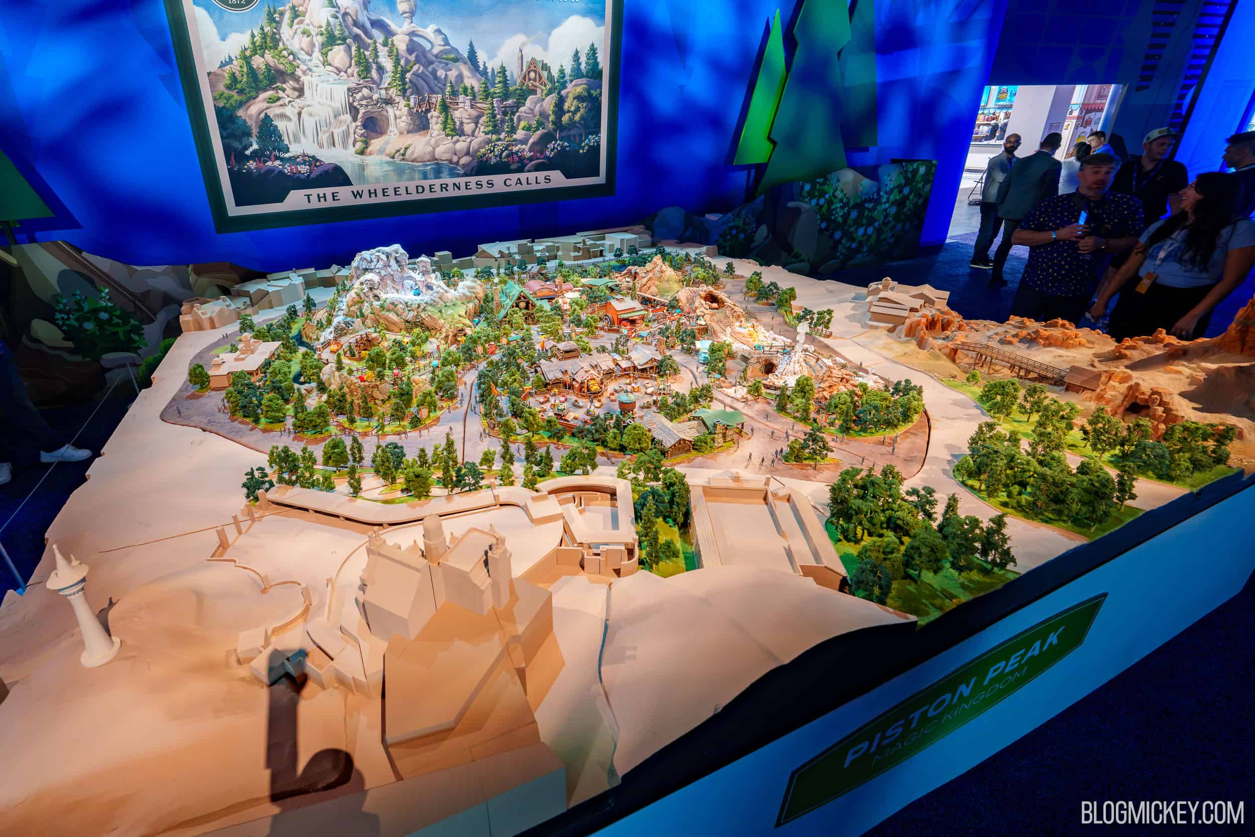 The full Piston Peak scale model on a show floor under blue lighting, with a mountain peak, forested areas and a green placard reading Piston Peak Magic Kingdom, beneath a poster reading The Wheelderness Calls