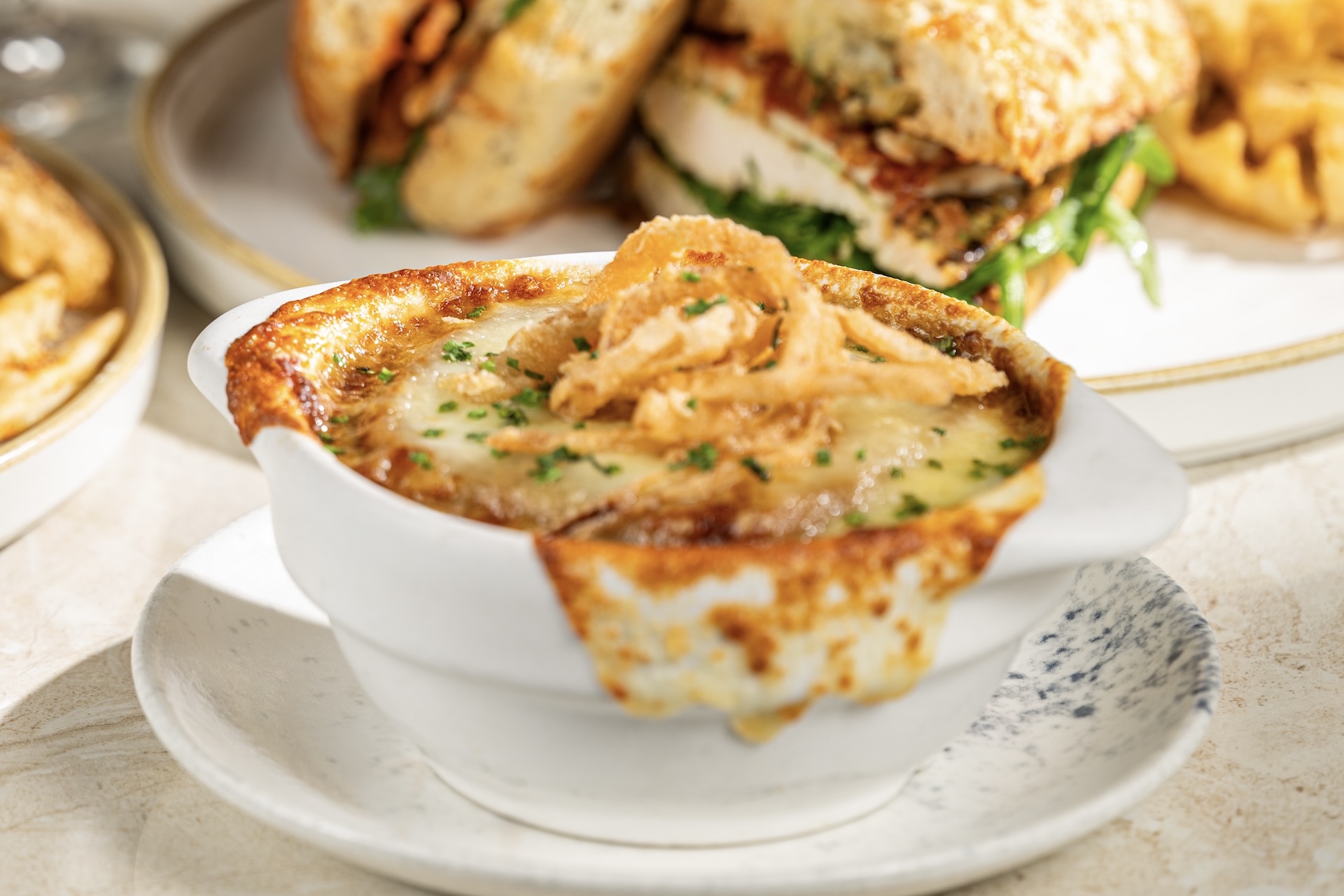 French Onion Soup in a white crock with browned cheese spilling over the rim and crispy onion straws on top, with a halved sandwich on a plate behind it