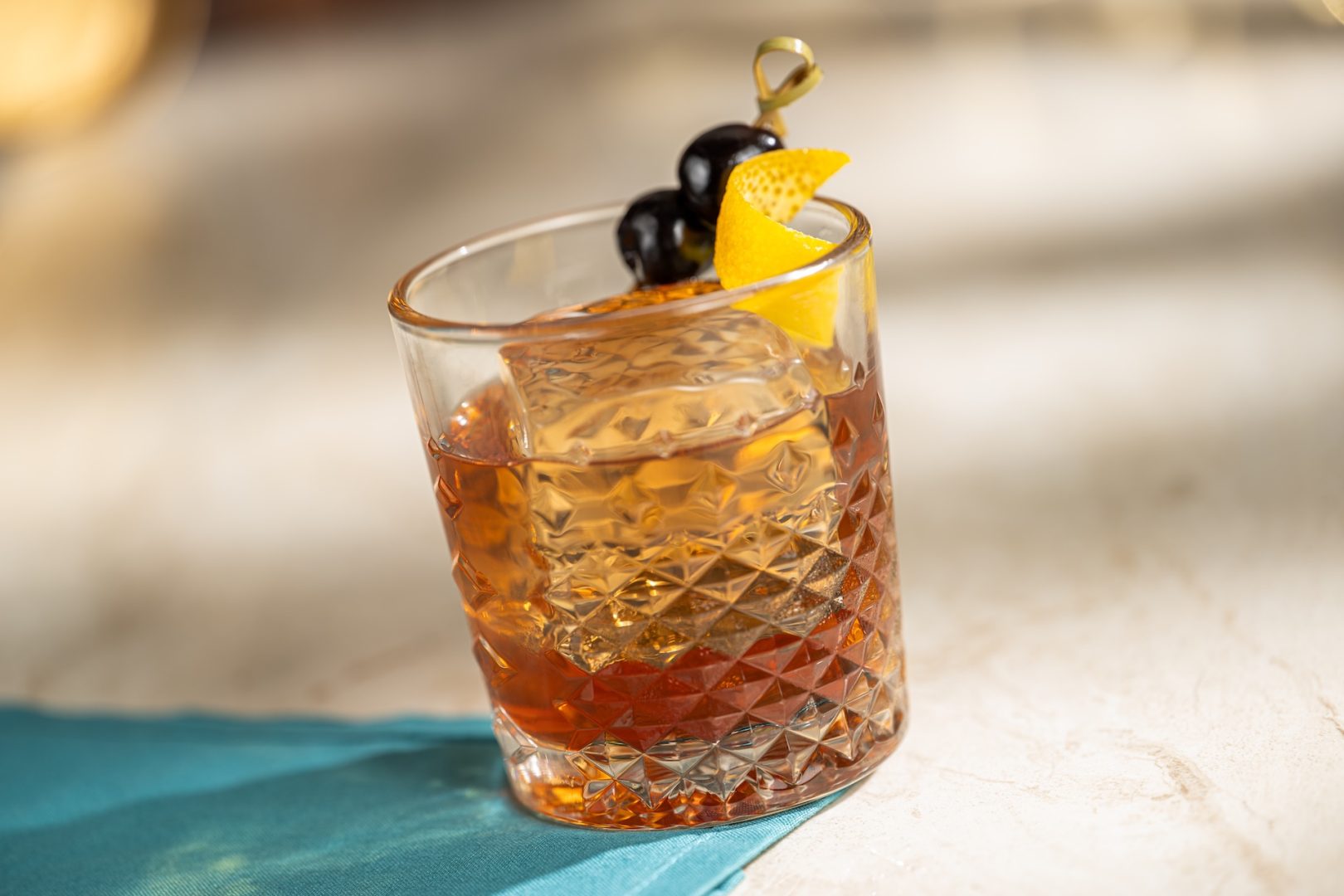 A New York Old Fashioned in a cut-crystal rocks glass over a large clear ice cube, garnished with two dark cherries and an orange twist on a gold pick