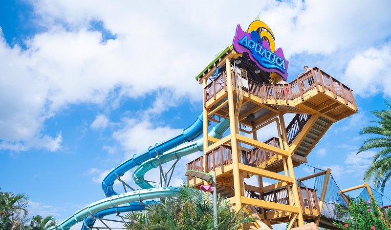 A tall wooden and yellow steel slide tower topped with the purple Aquatica logo, with blue and teal tube slides curling down from it under a bright blue sky with white clouds and palm fronds in the foreground