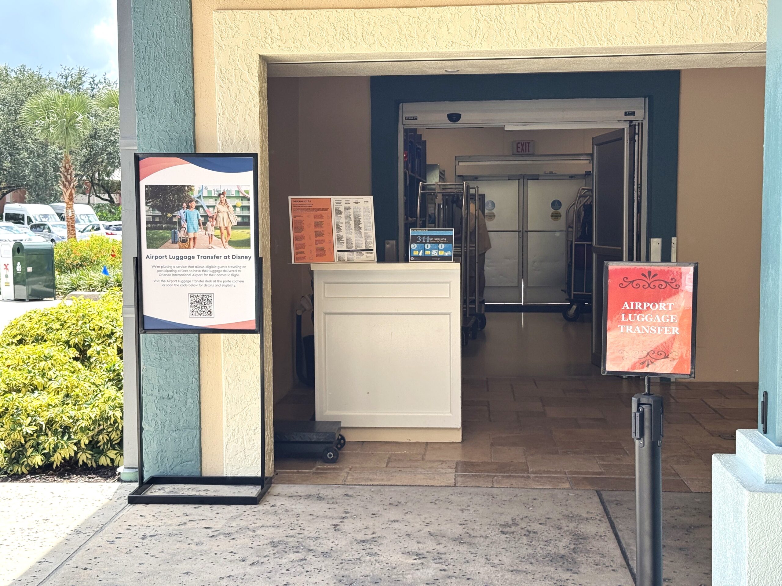 A resort porte cochere with a standing sign headed Airport Luggage Transfer at Disney showing a family walking with rolling suitcases and a QR code, a white podium beside it holding two TSA placards, one listing items that may not fly and one showing the 3-1-1 carry-on rules, a red stanchion sign reading AIRPORT LUGGAGE TRANSFER at the right, and metal bell carts standing in the vestibule behind
