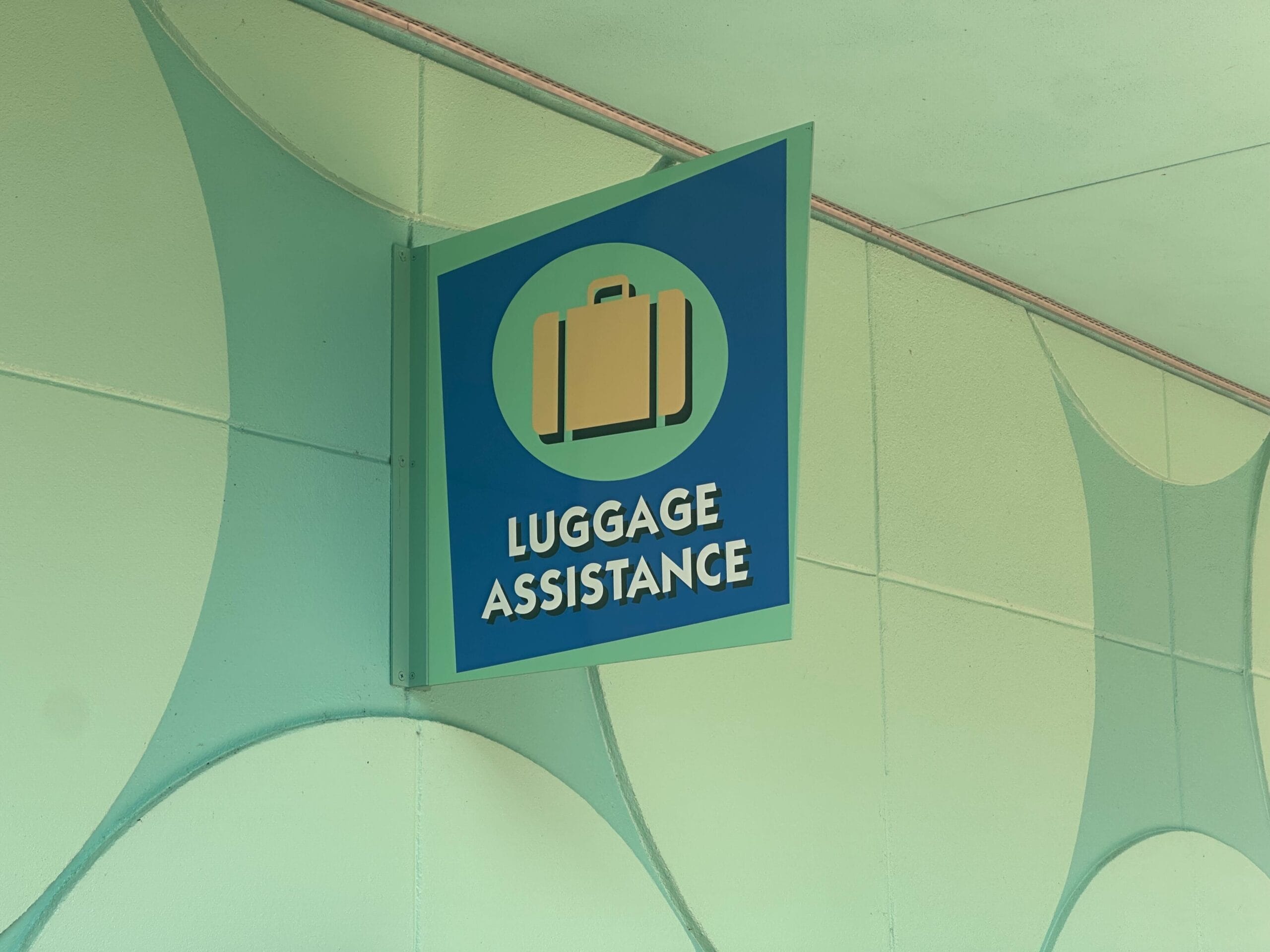 A projecting blade sign mounted on a pale green wall, dark blue with a green border, showing a tan suitcase inside a light green circle above the words LUGGAGE ASSISTANCE in white block letters