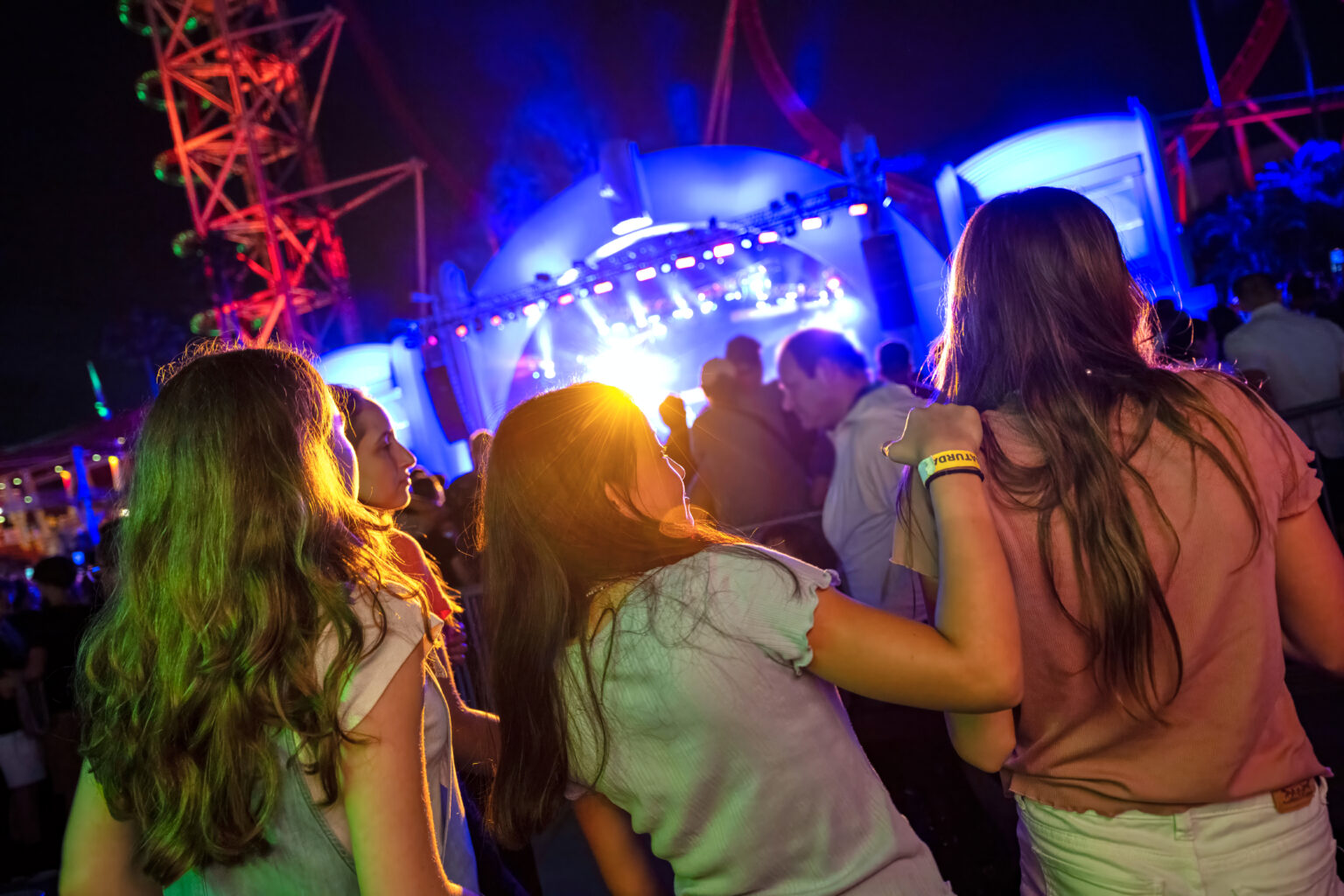 A night concert crowd photographed from behind, with a group of young women, one with an arm around another, facing a brightly blue lit outdoor stage, lighting rigs flaring above it, and the red steel structure of the Hollywood Rip Ride Rockit coaster rising at the left edge against a black sky